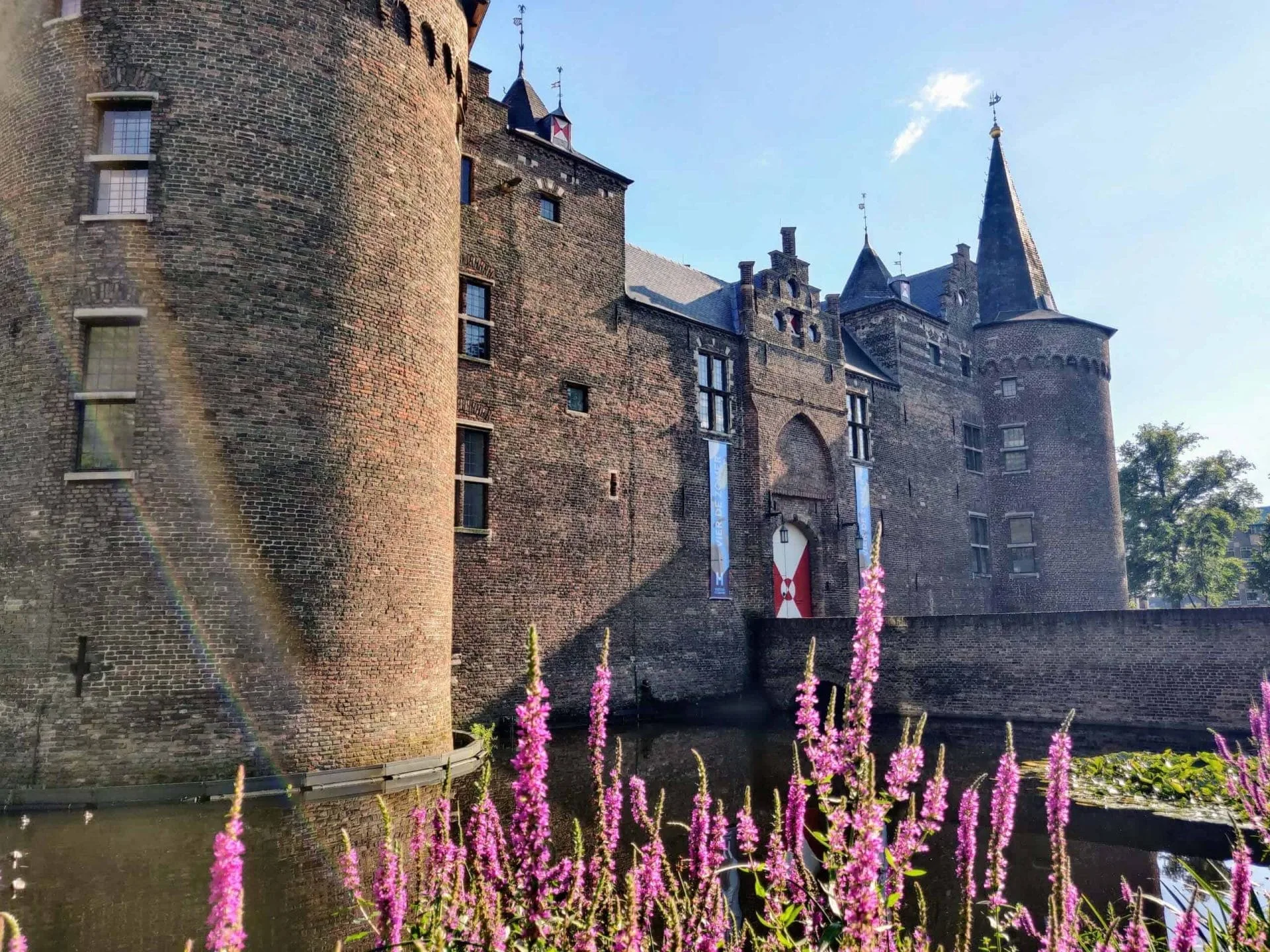 Zijaanzicht van het middeleeuwse Kasteel Helmond met ronde torens en een slotgracht, omgeven door paarse bloemen