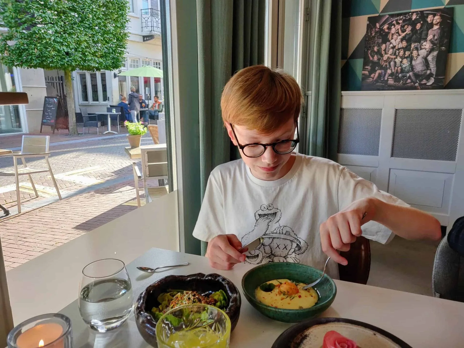 Een jongen met een bril zit aan een tafel in een restaurant en snijdt met mes en vork in een verfijnd gerecht