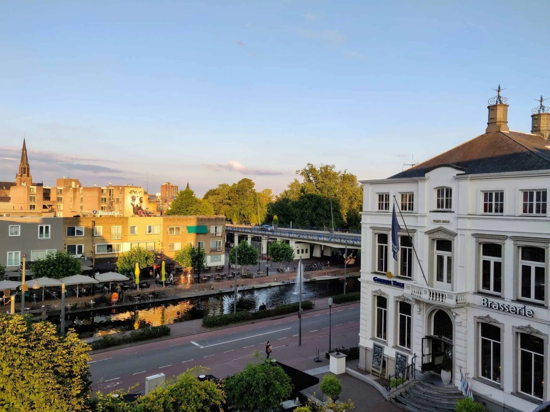 Uitzicht vanuit Hotel West-Ende op een kanaal met terrassen, een brug en de Lambertuskerk in Helmond bij zonsondergang