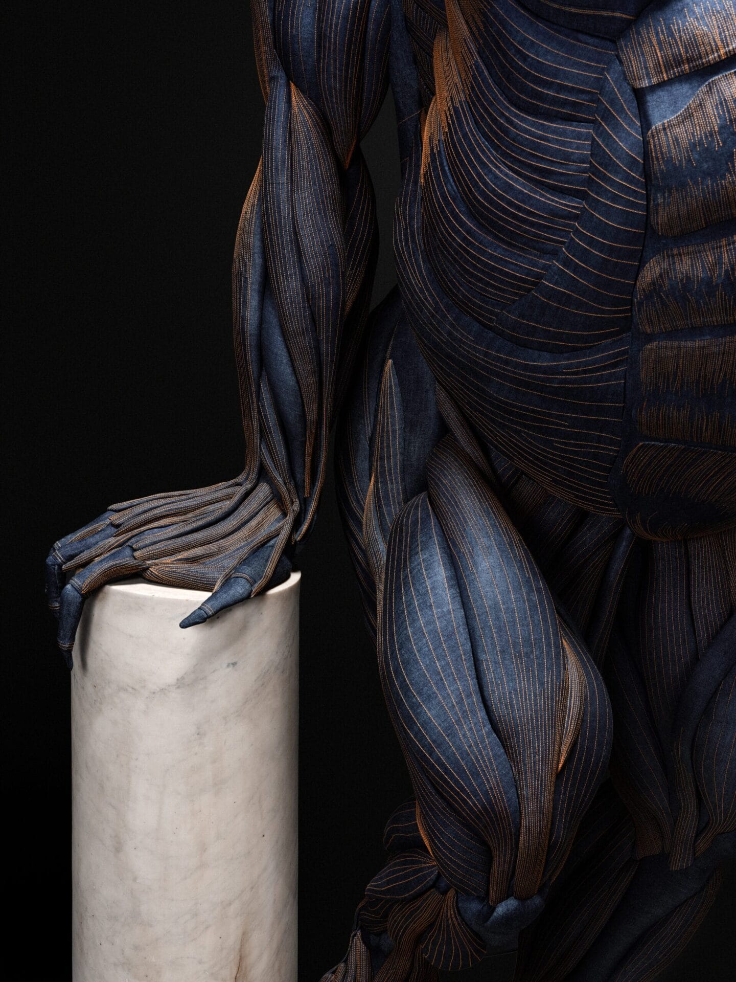 Detail van de Denim Gorilla met zichtbare spieren van handgestikt denim, rustend met een hand op een marmeren zuil