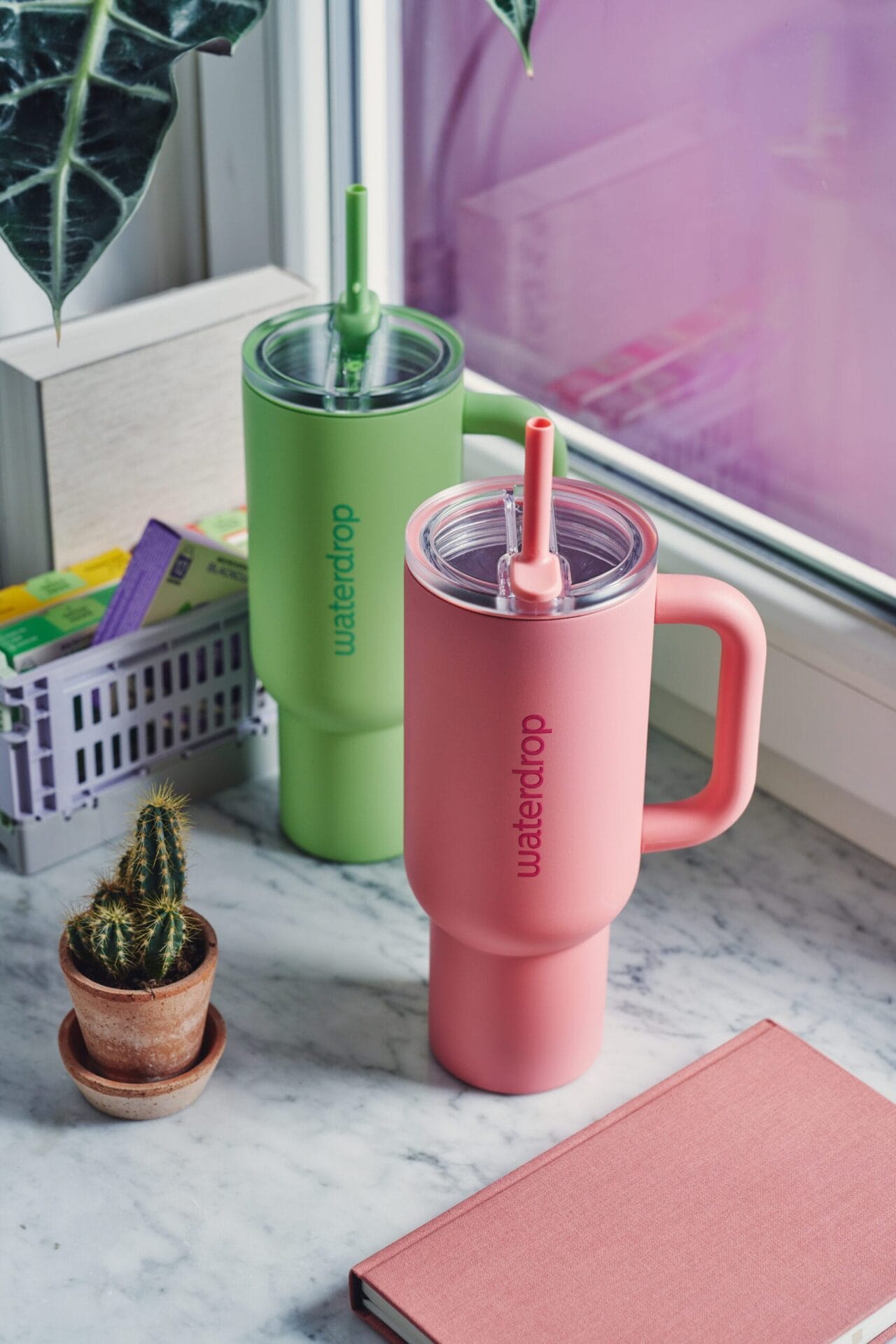 Een roze en een groene Waterdrop Explorer Thermo Drinkbeker met handvat en rietje op een marmeren vensterbank
