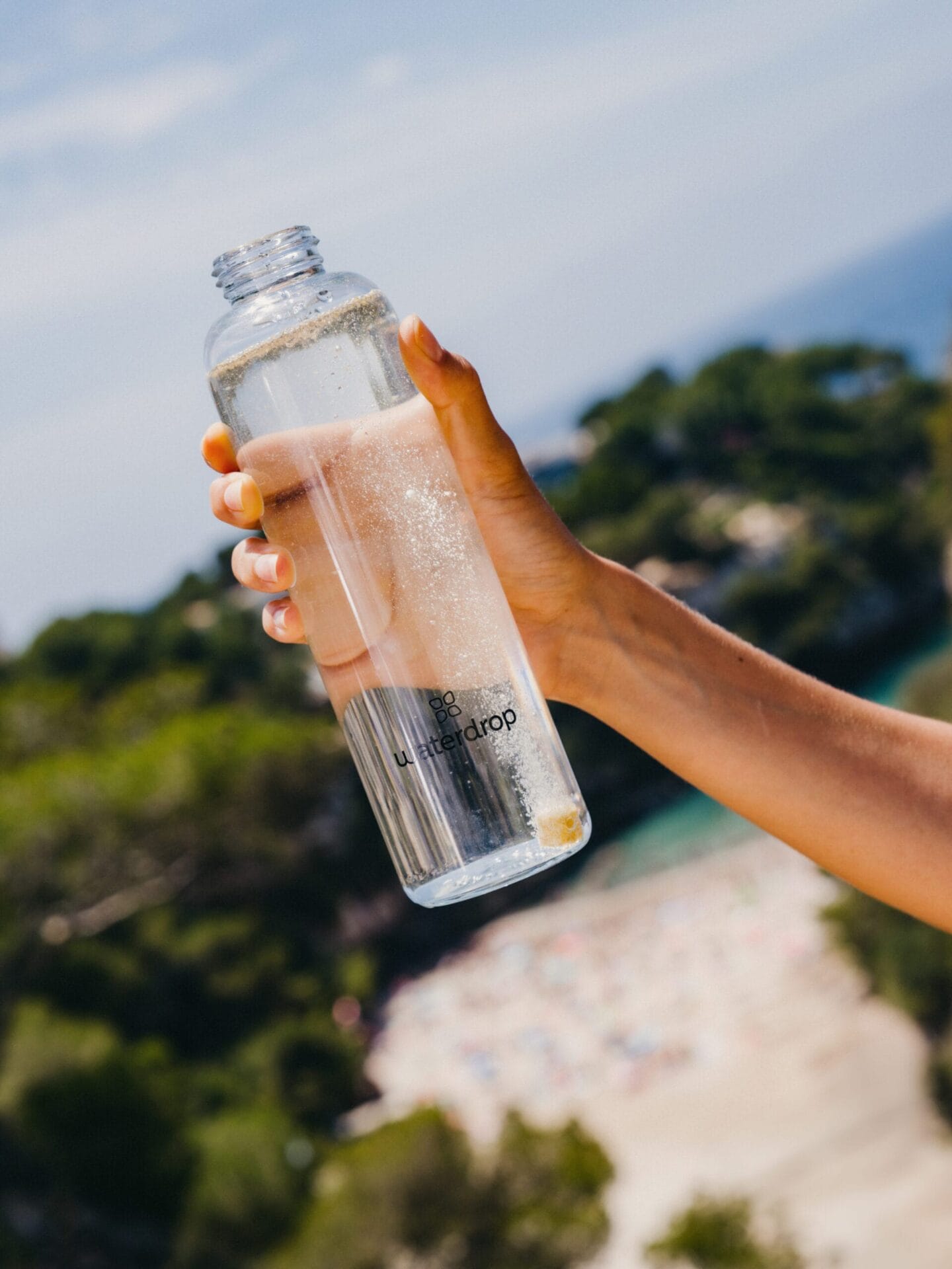 Een hand houdt een glazen Waterdrop-fles vast waarin een bruisblokje in het water oplost met een zonnig strand op de achtergrond
