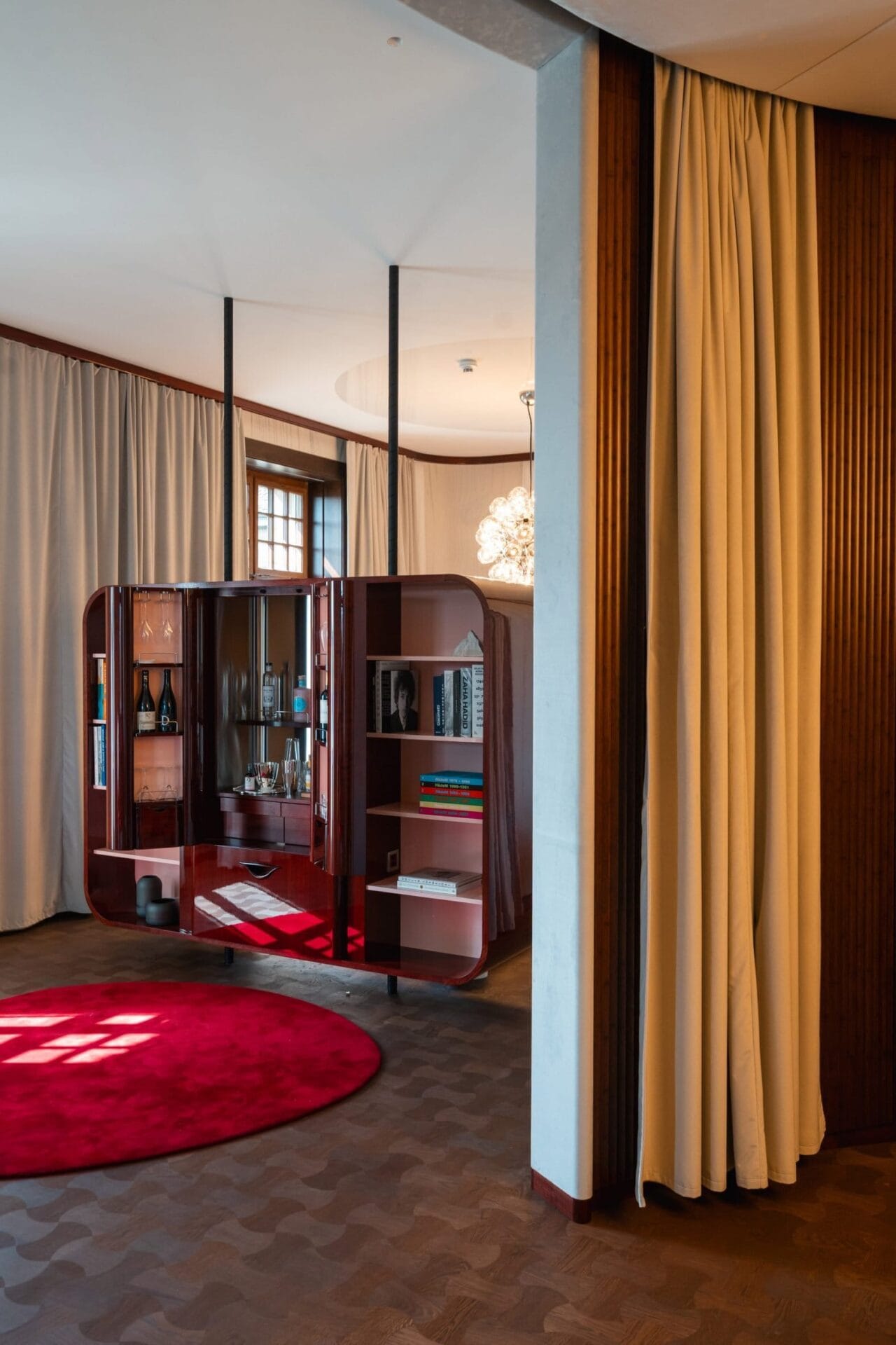 Modern interieur van een suite met een mahoniekleurig kastmeubel, een rond rood vloerkleed en kamerhoge gordijnen