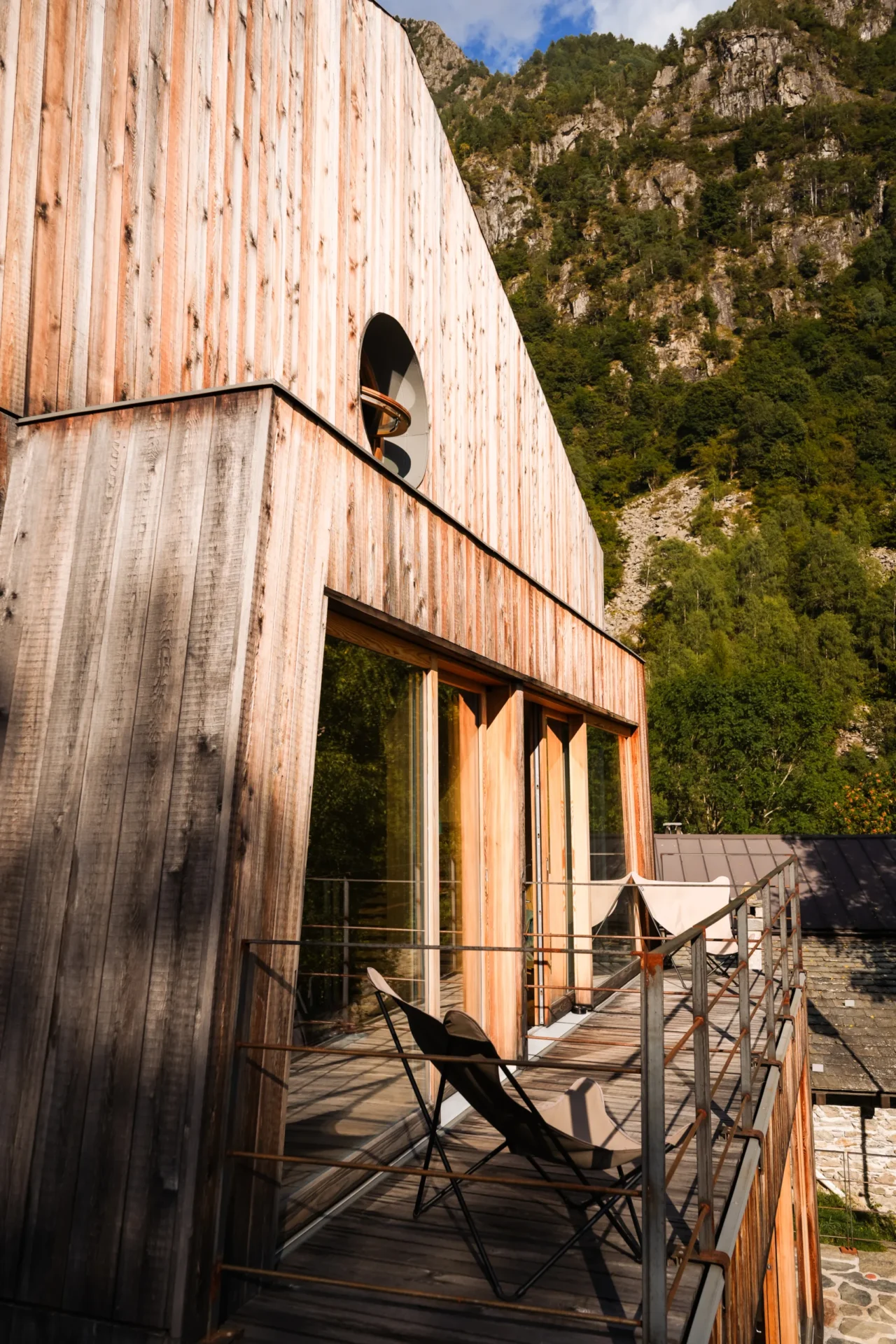 Modern houten gebouw van Rifugio Val di Togno met een terras en ligstoelen tegen een beboste bergwand
