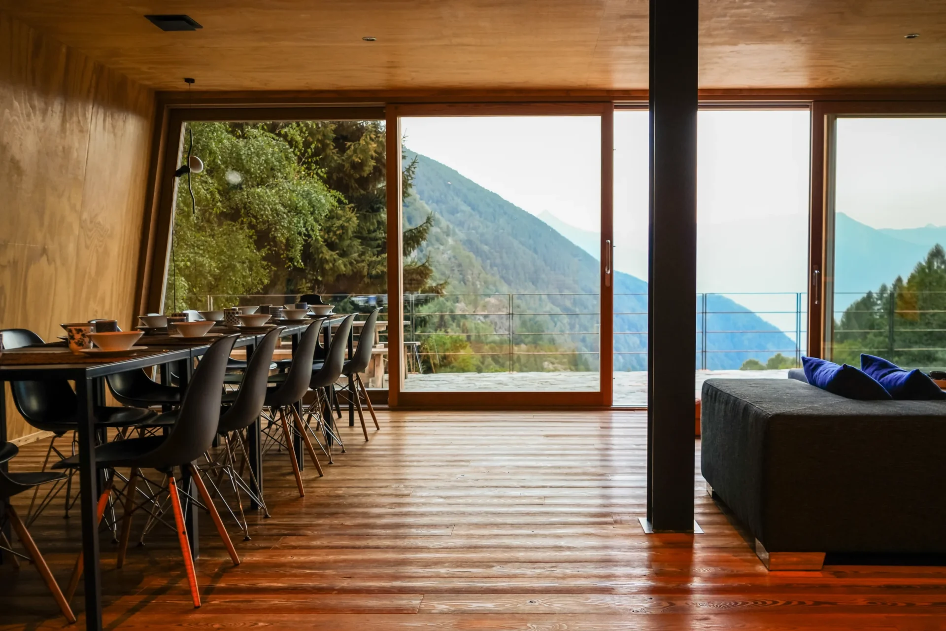 Modern interieur van Rifugio Val di Togno met een gedekte eettafel en uitzicht op de beboste bergen door grote ramen