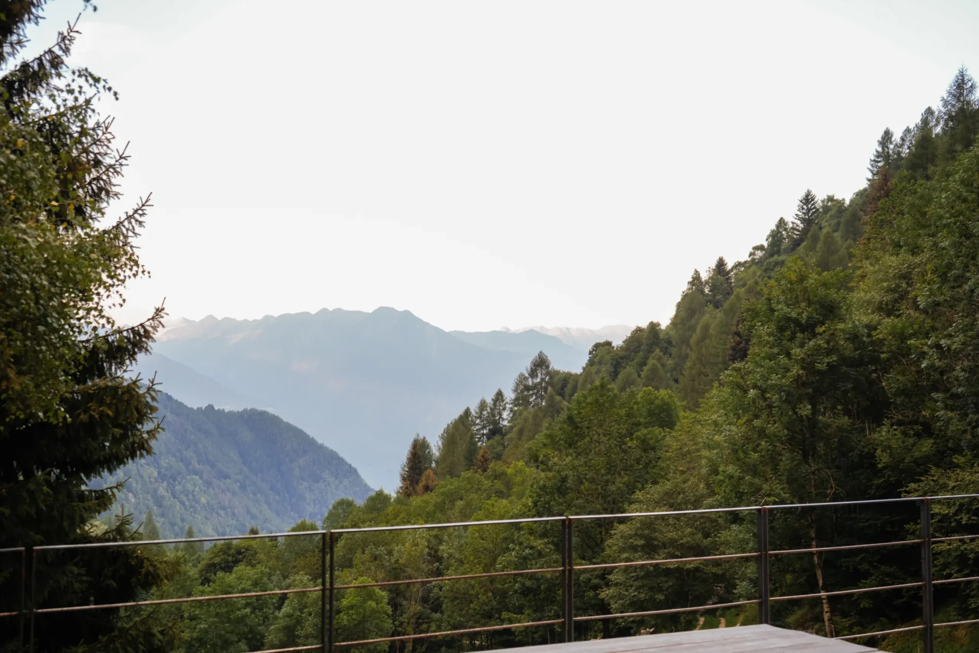 Uitzicht vanaf een terras van Rifugio Val di Togno op beboste bergen en verre bergtoppen in de nevel