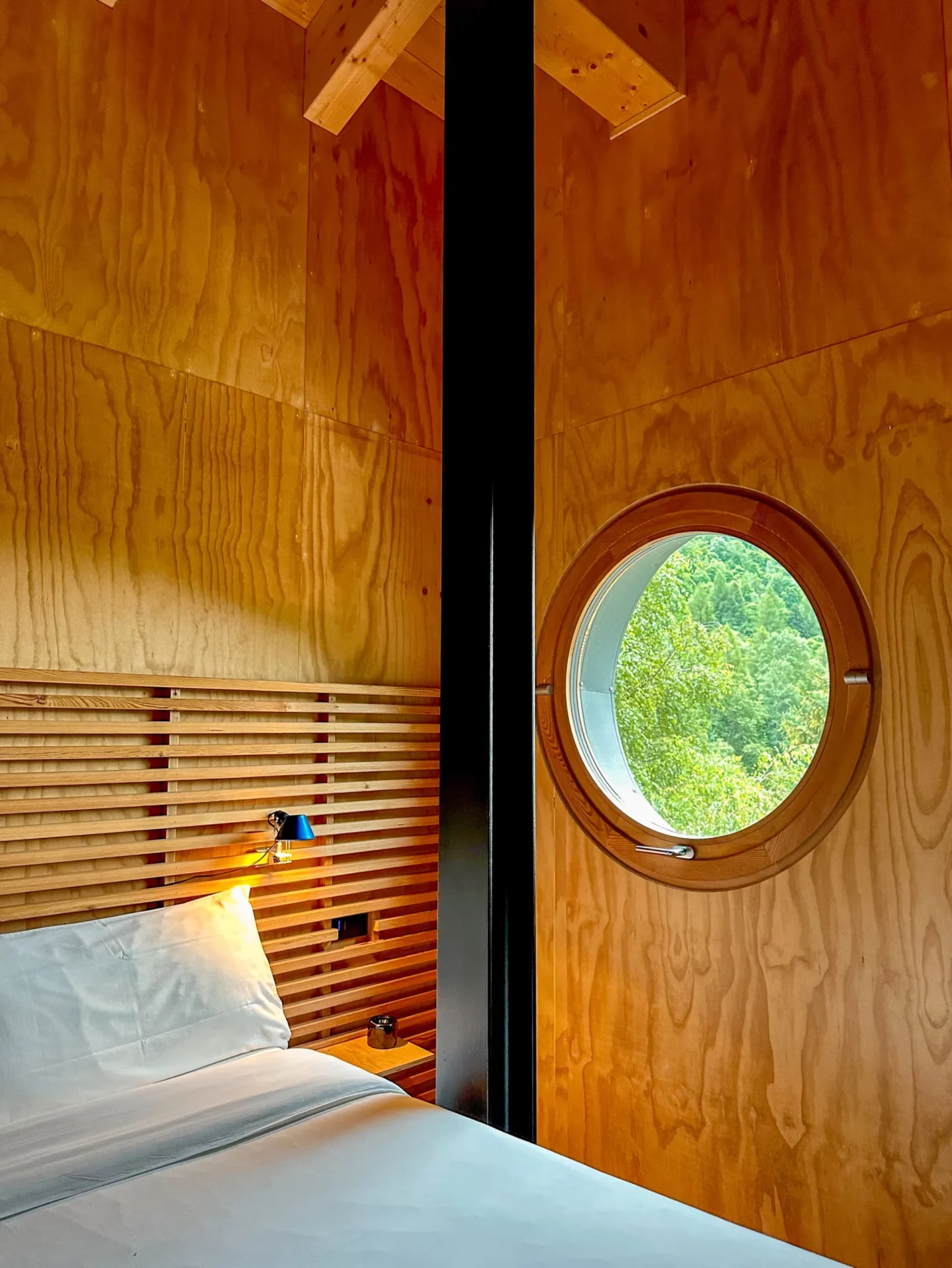 Slaapkamer in Rifugio Val di Togno met houten wanden, een rond raam met uitzicht op bos en een bed met houten hoofdbord