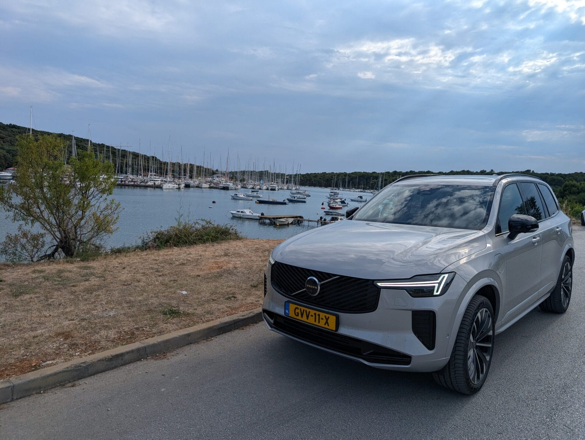 Grijze Volvo XC90 T8 geparkeerd langs de kust in Kroatië met een jachthaven vol zeilboten op de achtergrond