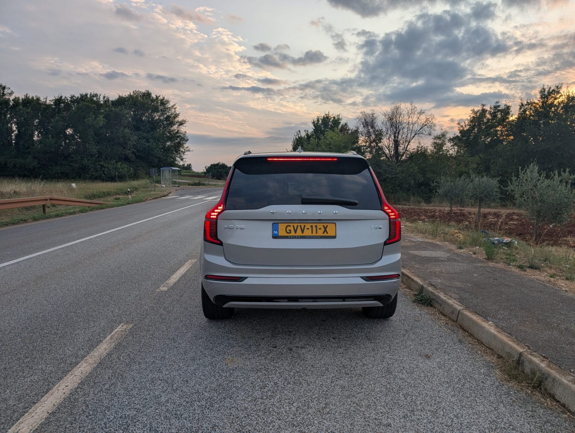 Achteraanzicht van een zilveren Volvo XC90 T8 op een weg tijdens zonsondergang in een heuvelachtig landschap