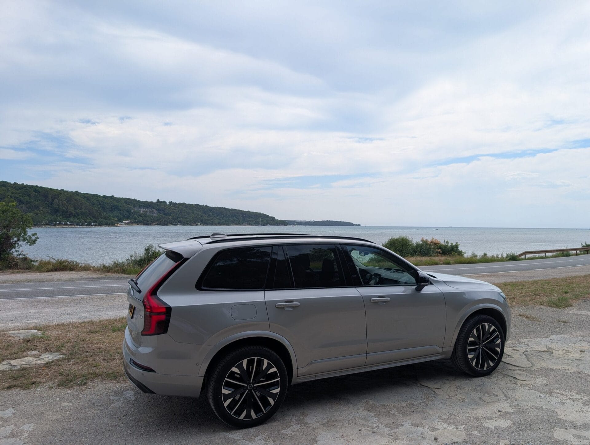 Zijaanzicht van een grijze Volvo XC90 geparkeerd langs een kustweg in Kroatië met uitzicht op de zee en beboste heuvels