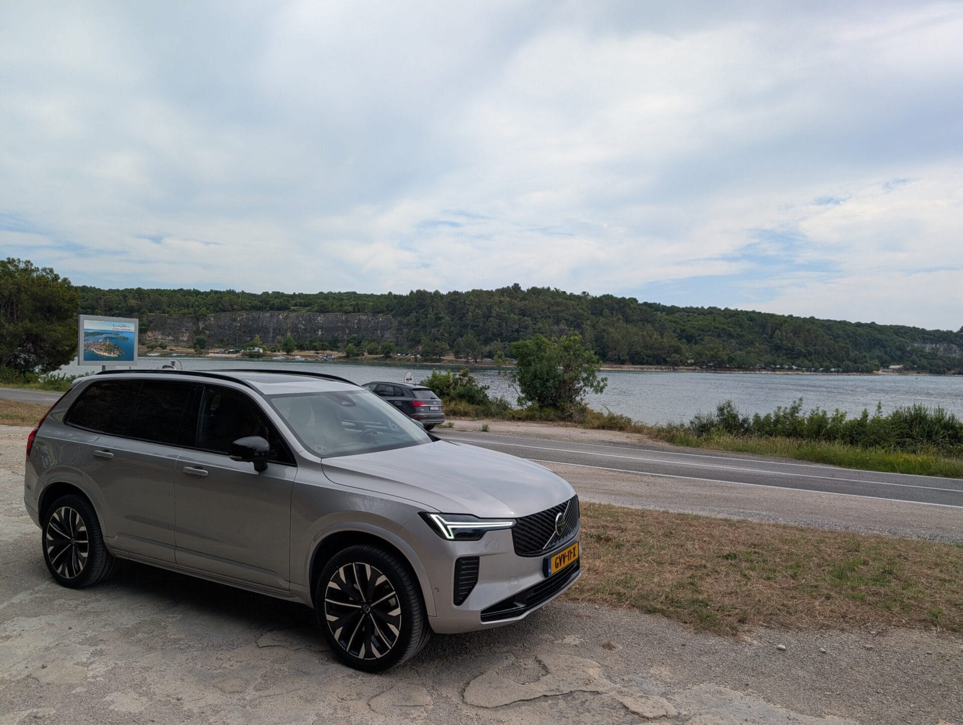 Zijaanzicht van een zilveren Volvo XC90 geparkeerd langs een kustweg in Kroatië met uitzicht op het water en beboste heuvels