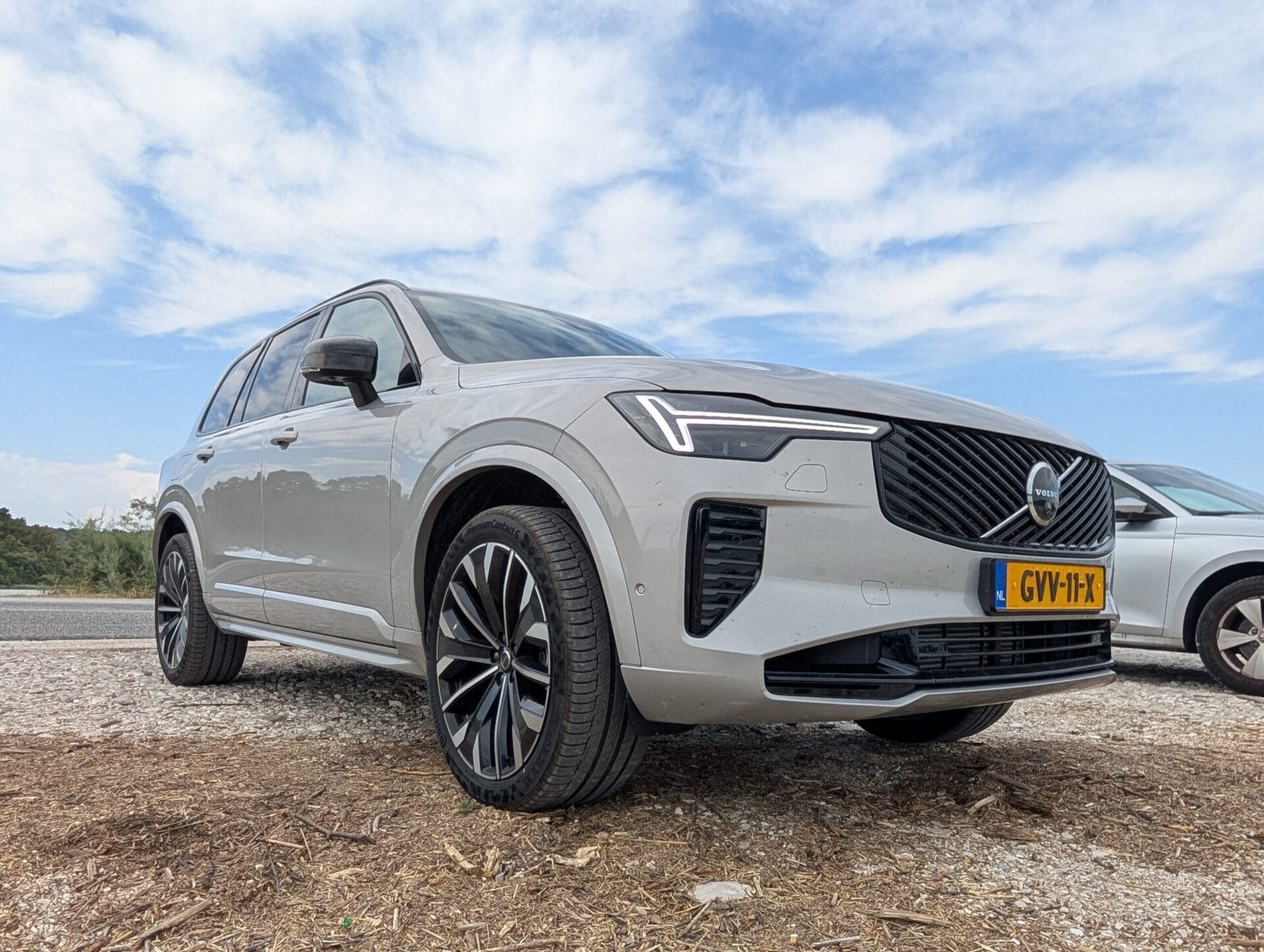 Vooraanzicht van een grijze Volvo XC90 T8 geparkeerd op een onverharde ondergrond onder een lichtbewolkte blauwe lucht