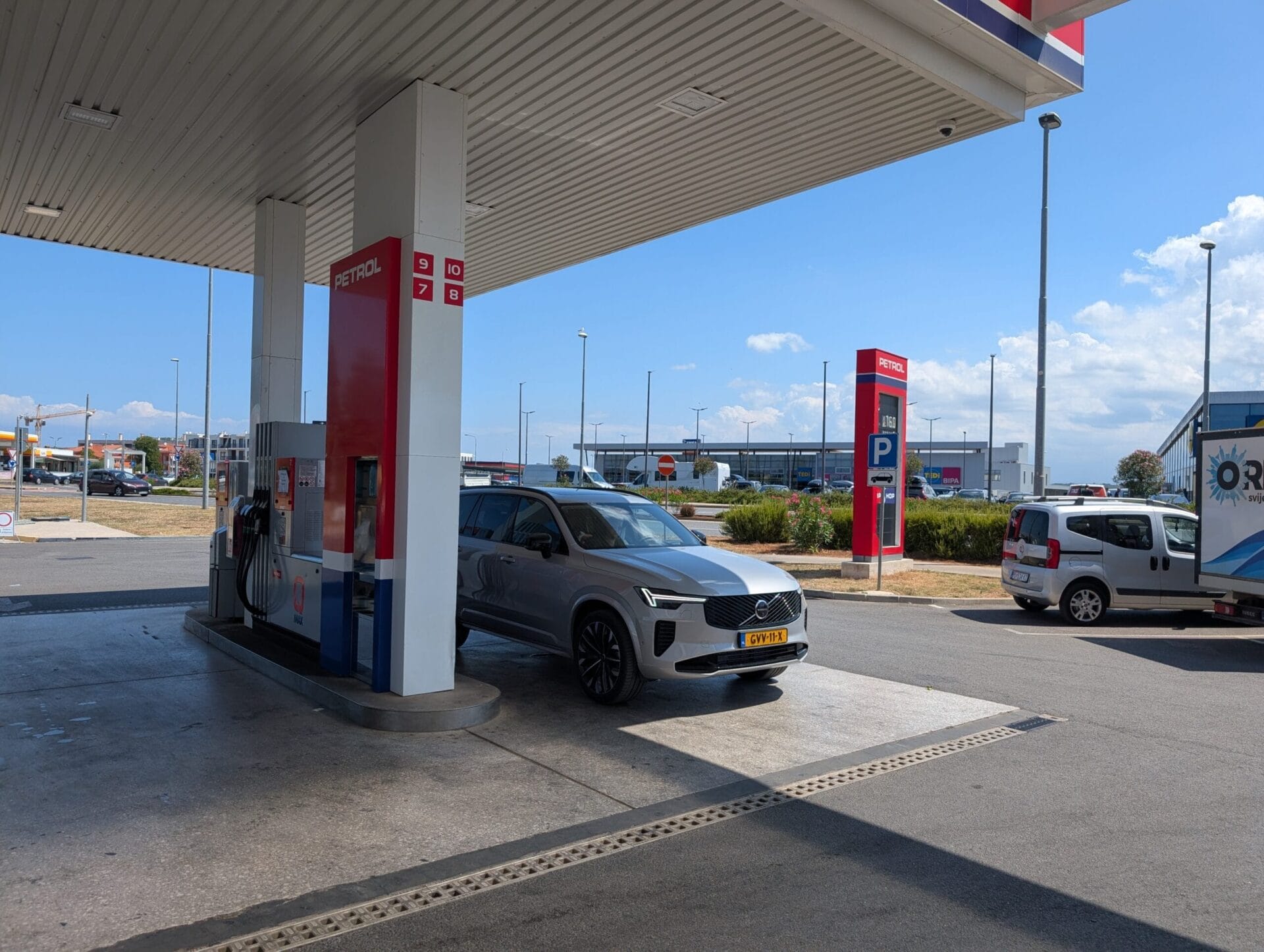 Een zilveren Volvo XC90 T8 staat geparkeerd bij een Petrol-tankstation onder een overkapping tijdens een zonnige dag