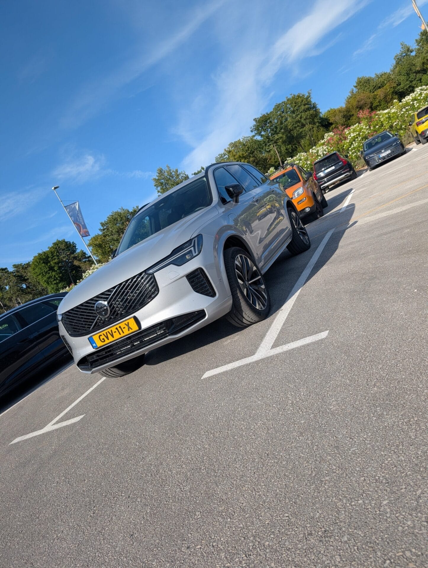 Vooraanzicht van een zilveren Volvo XC90 geparkeerd op een parkeerplaats onder een blauwe lucht