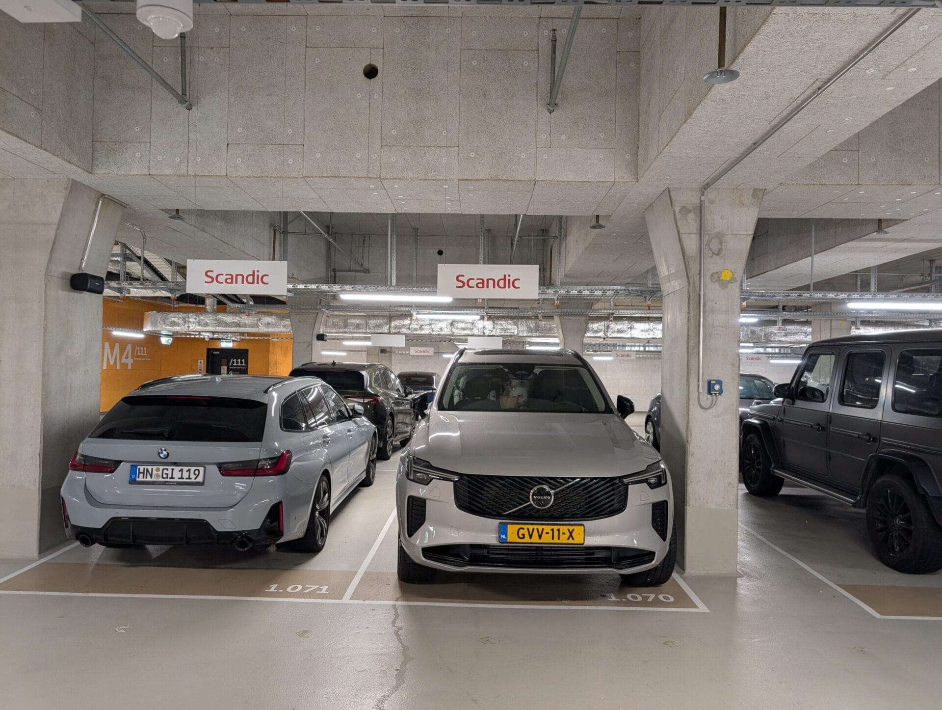 Een zilveren Volvo XC90 geparkeerd in een parkeergarage van een Scandic hotel naast een grijze BMW
