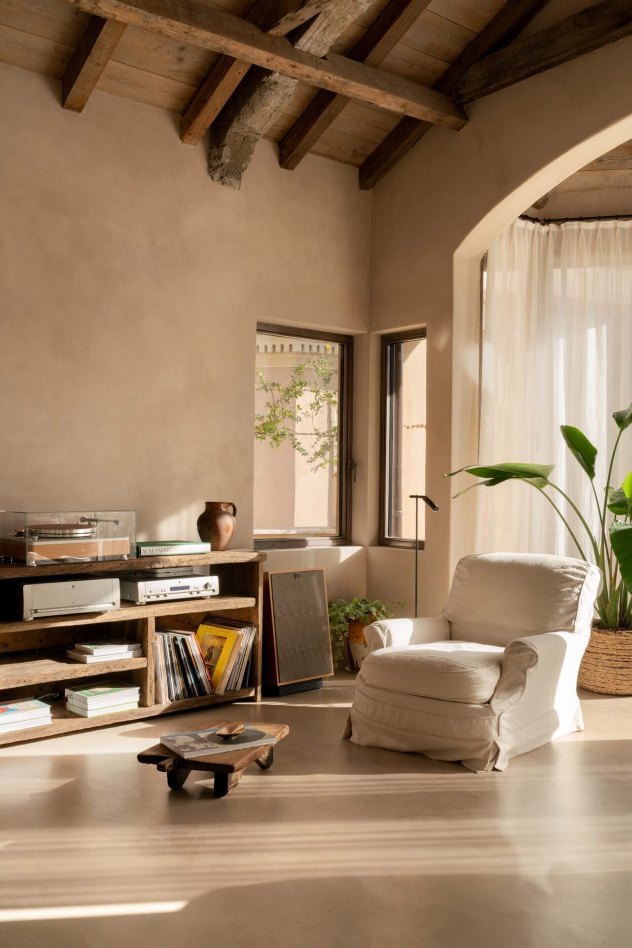 Een minimalistisch interieur met kalkmuren, houten plafondbalken, een rustieke kast met platenspeler en een witte fauteuil