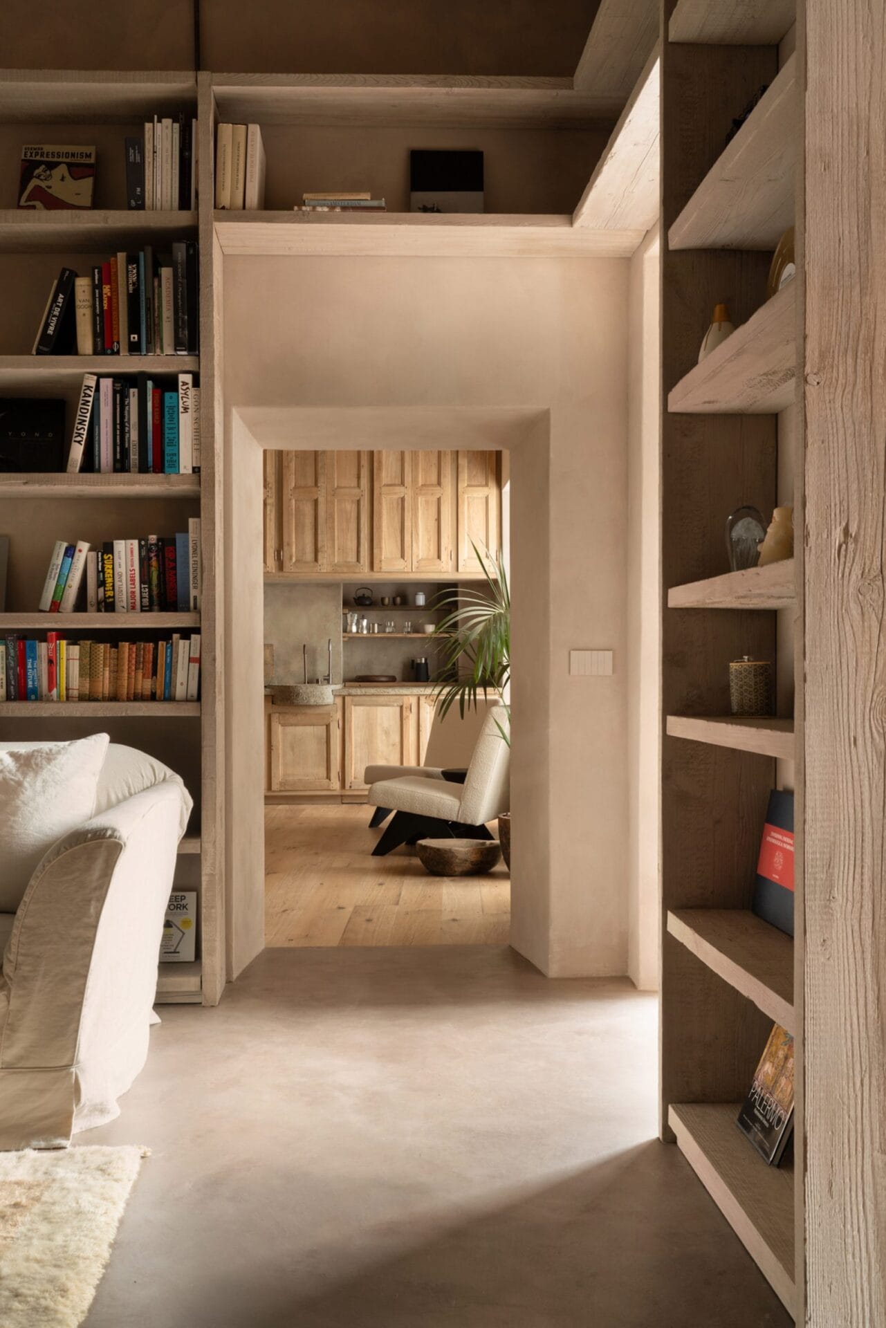 Een doorkijk vanuit een woonkamer met houten boekenkasten naar een lichte keuken met houten kasten en witte fauteuils