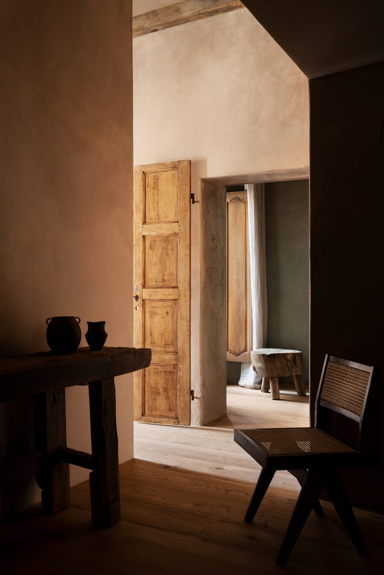 Een minimalistisch interieur in wabi-sabi stijl met ruwe kalkmuren, een houten tafel, een rieten stoel en antieke deuren
