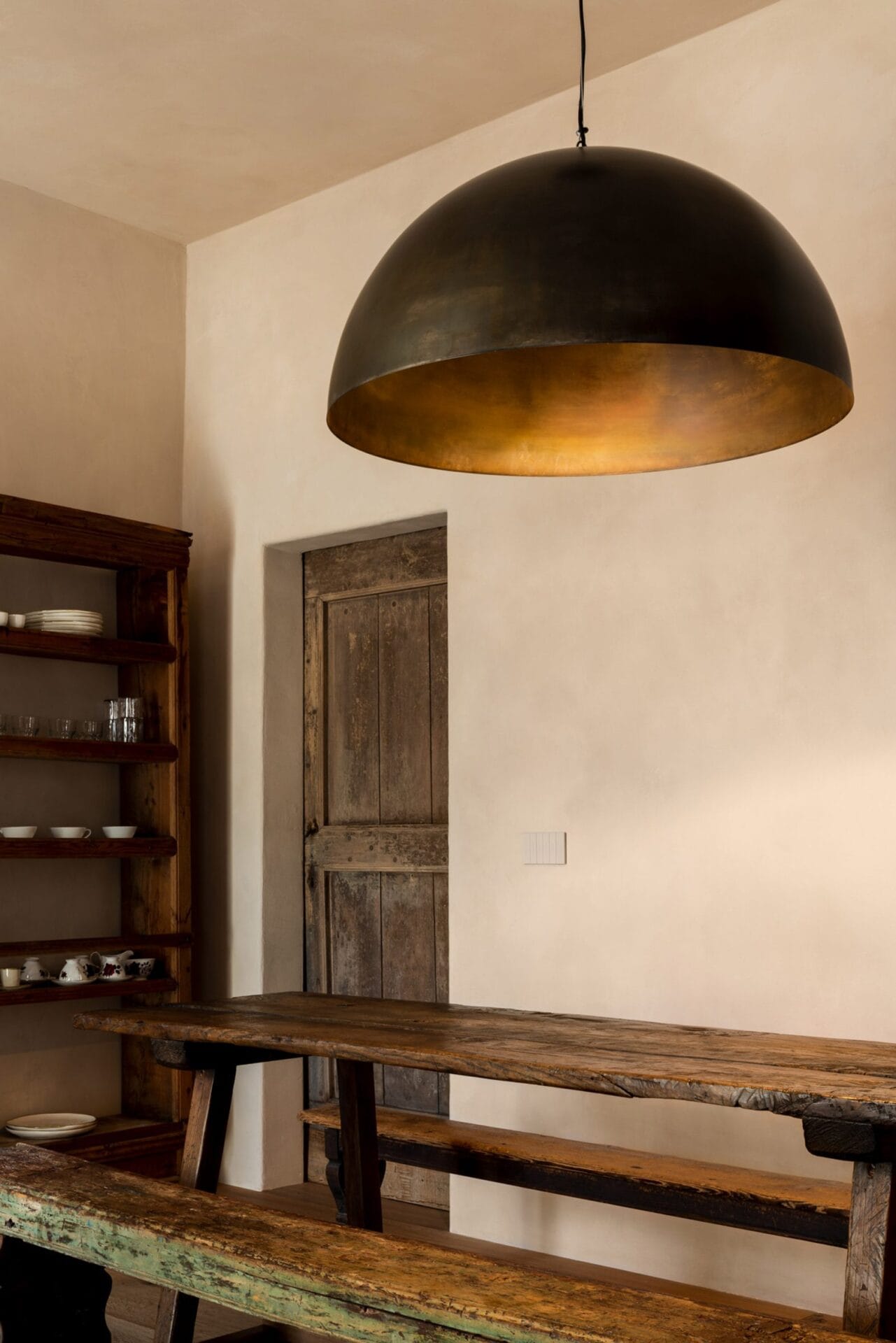 Een minimalistisch interieur met een robuuste houten tafel, banken, een antieke deur en een grote koepelvormige hanglamp
