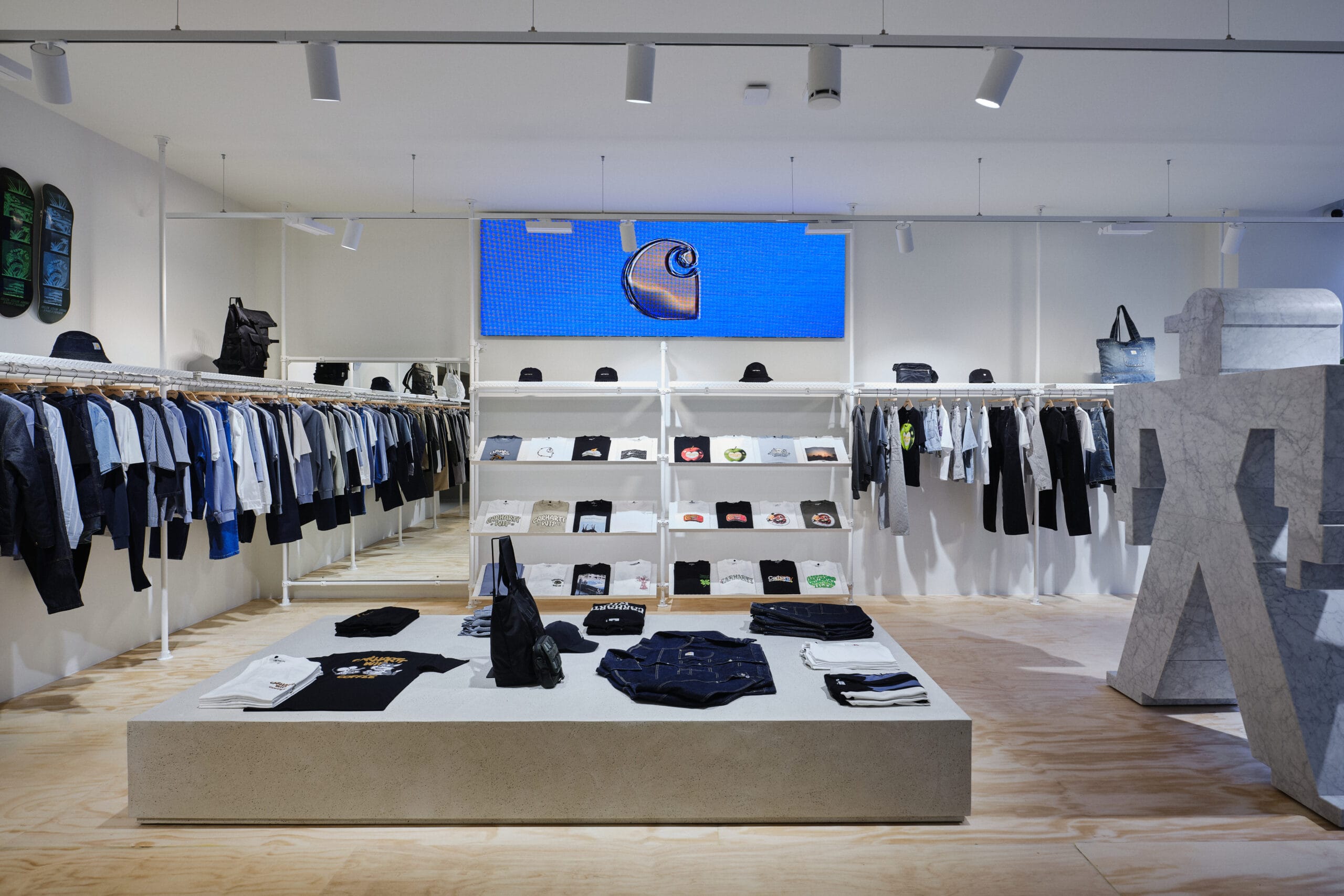 Interieur van een moderne Carhartt WIP kledingwinkel met minimalistische witte rekken en een groot blauw digitaal scherm