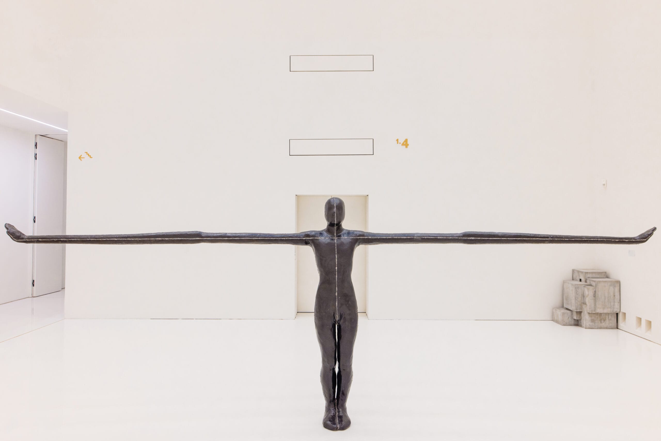 Een donker, mensvormig standbeeld met extreem lange, horizontaal uitgestrekte armen in een witte museumzaal