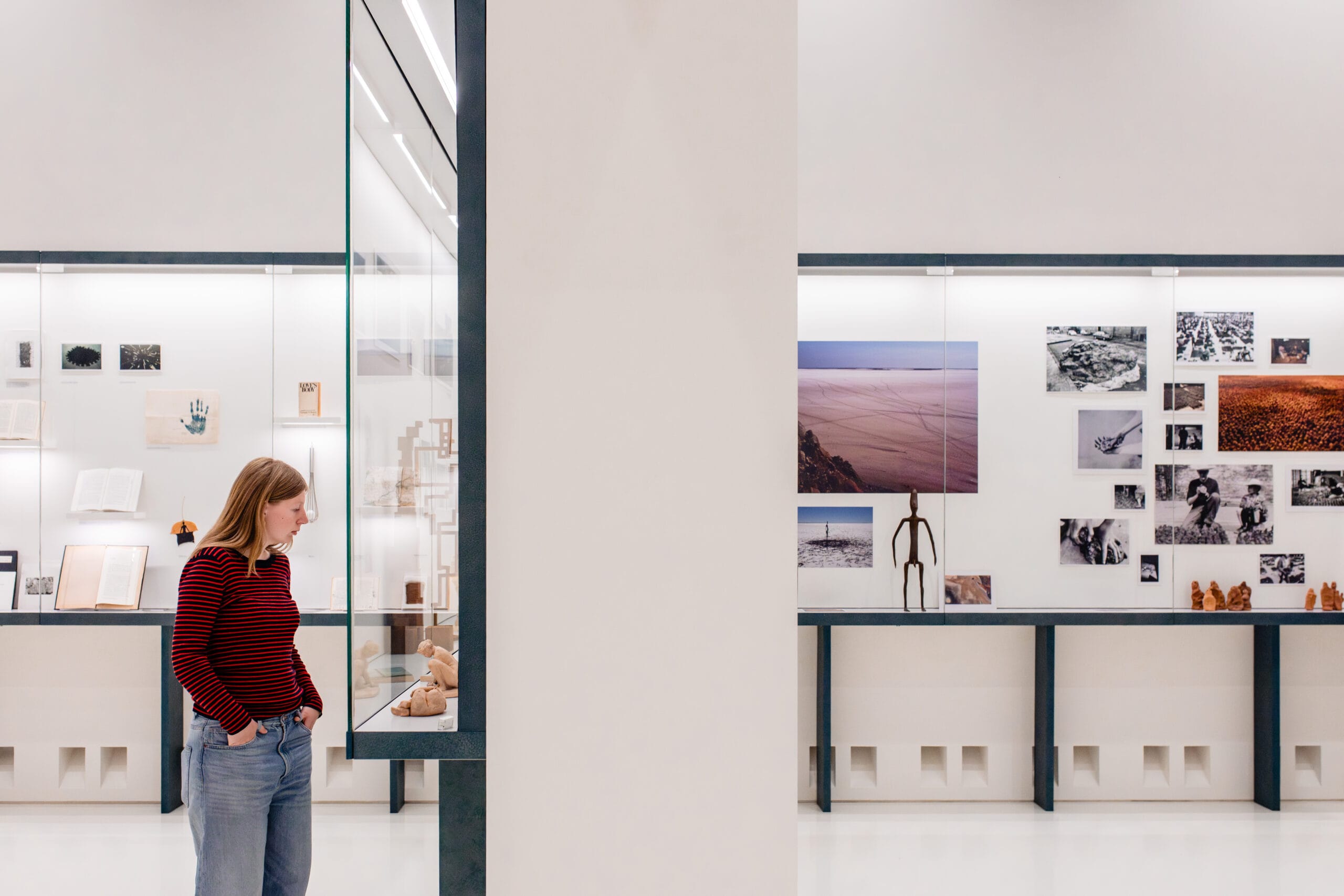Een vrouw in een rood-zwart gestreepte trui bekijkt kunstwerken in een modern, licht museum met glazen vitrines