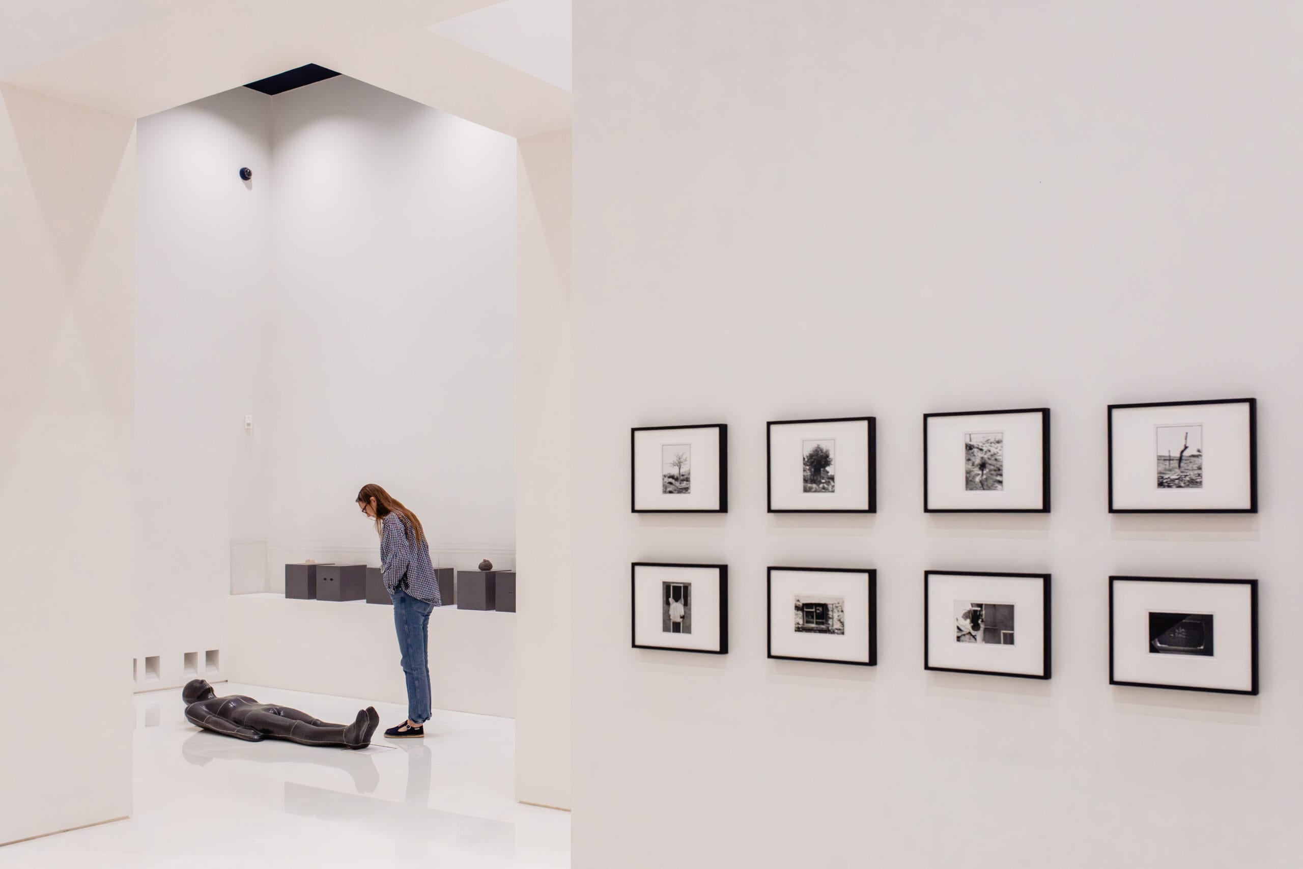 Een bezoeker in een wit verlichte museumzaal kijkt naar een liggend standbeeld op de grond naast een wand met fotolijsten
