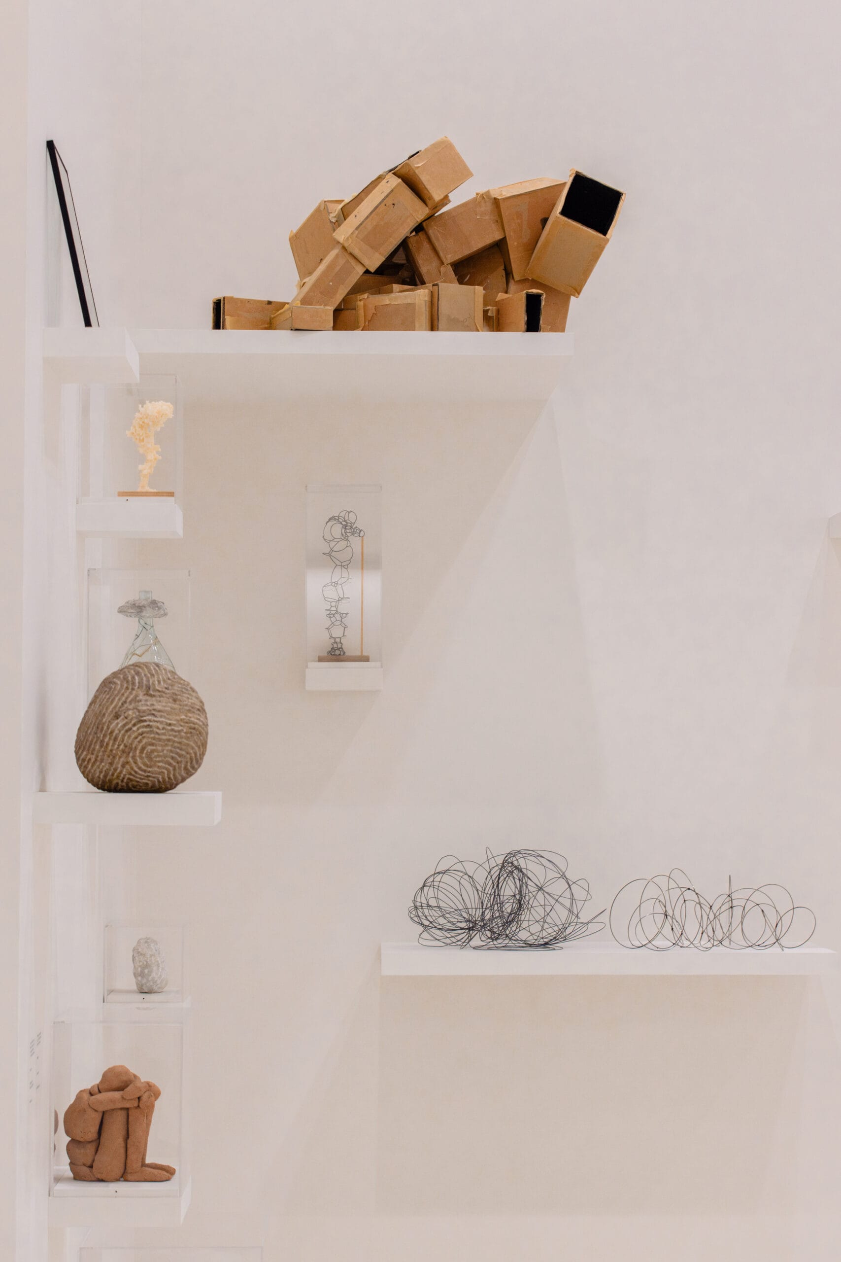 Diverse abstracte sculpturen van karton, draad en steen tentoongesteld op witte planken tegen een witte muur