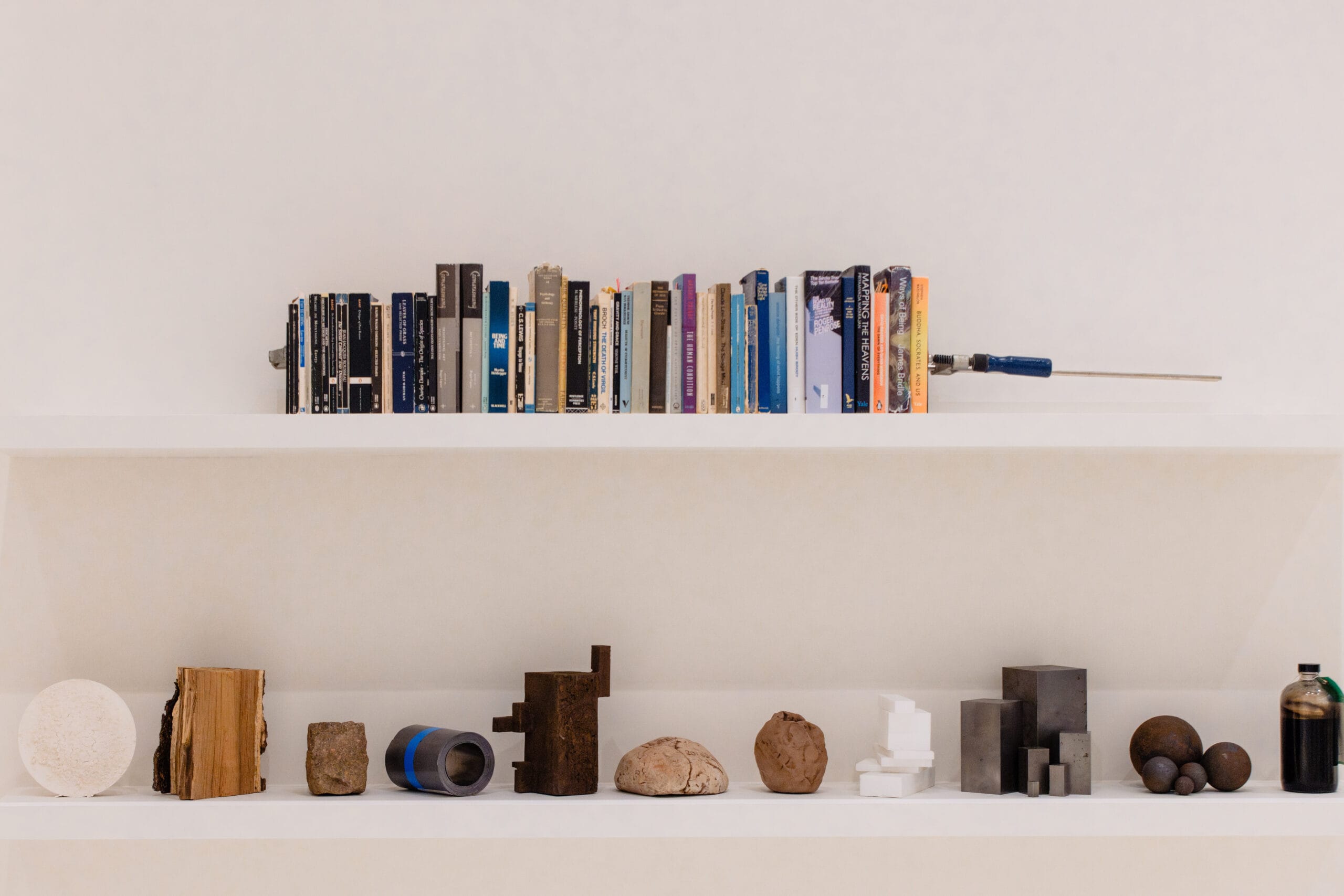 Twee witte planken tegen een witte muur met boeken, een lijmklem en diverse abstracte objecten van hout, steen en metaal