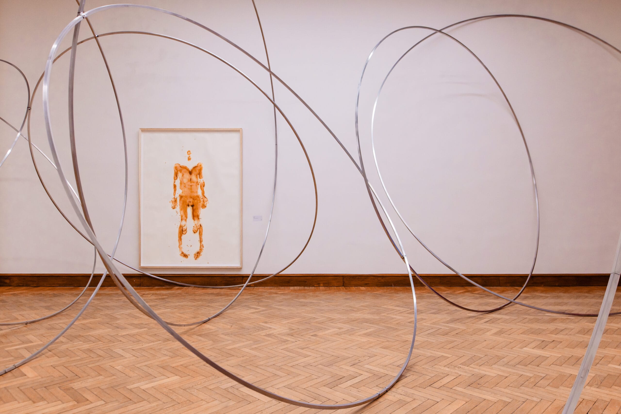 Grote metalen ringen in een museumzaal voor een schilderij van een menselijk figuur in bruintinten