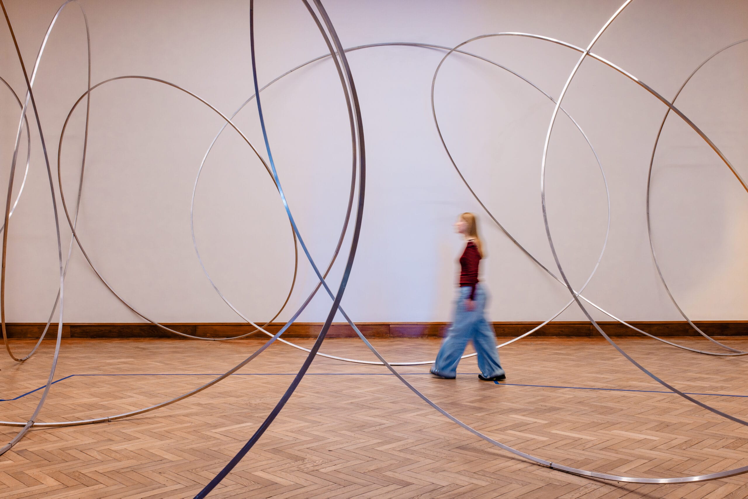 Een persoon loopt langs een kunstinstallatie van grote, dunne metalen ringen in een museumzaal met een houten vloer
