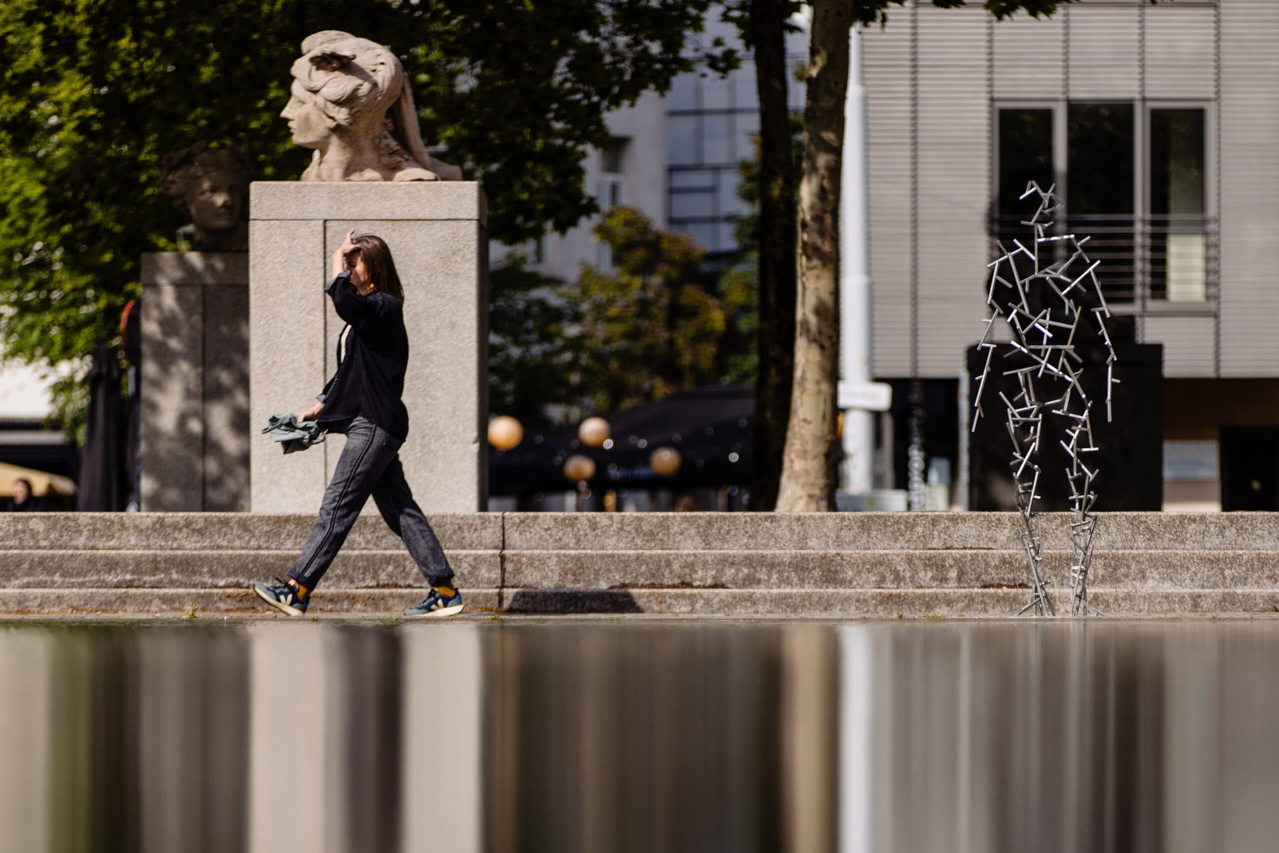Een vrouw loopt langs een modern metalen standbeeld en klassieke bustes op een stadsplein