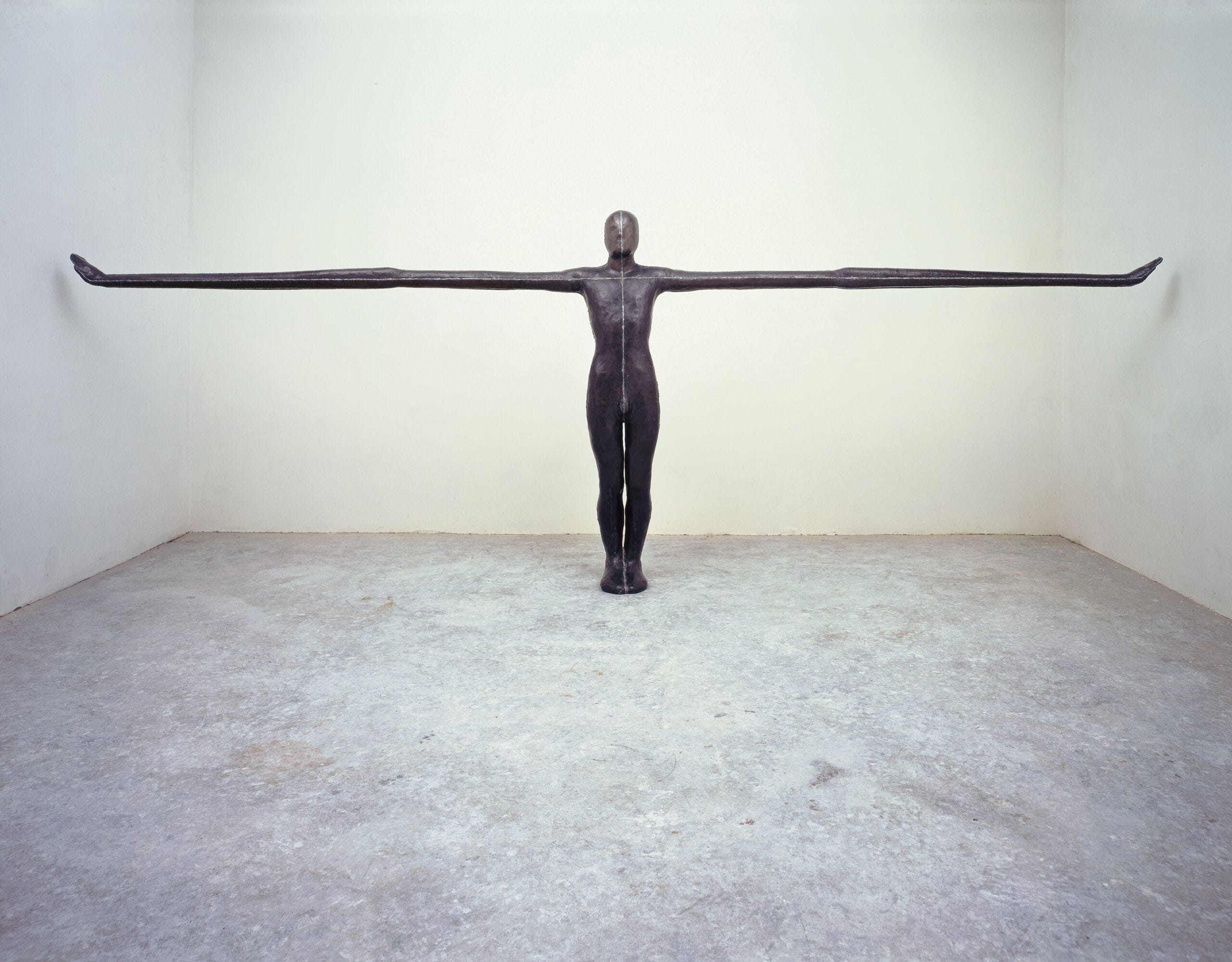 Een donker metalen sculptuur van een menselijke figuur met extreem lange, horizontaal uitgestrekte armen in een witte kamer