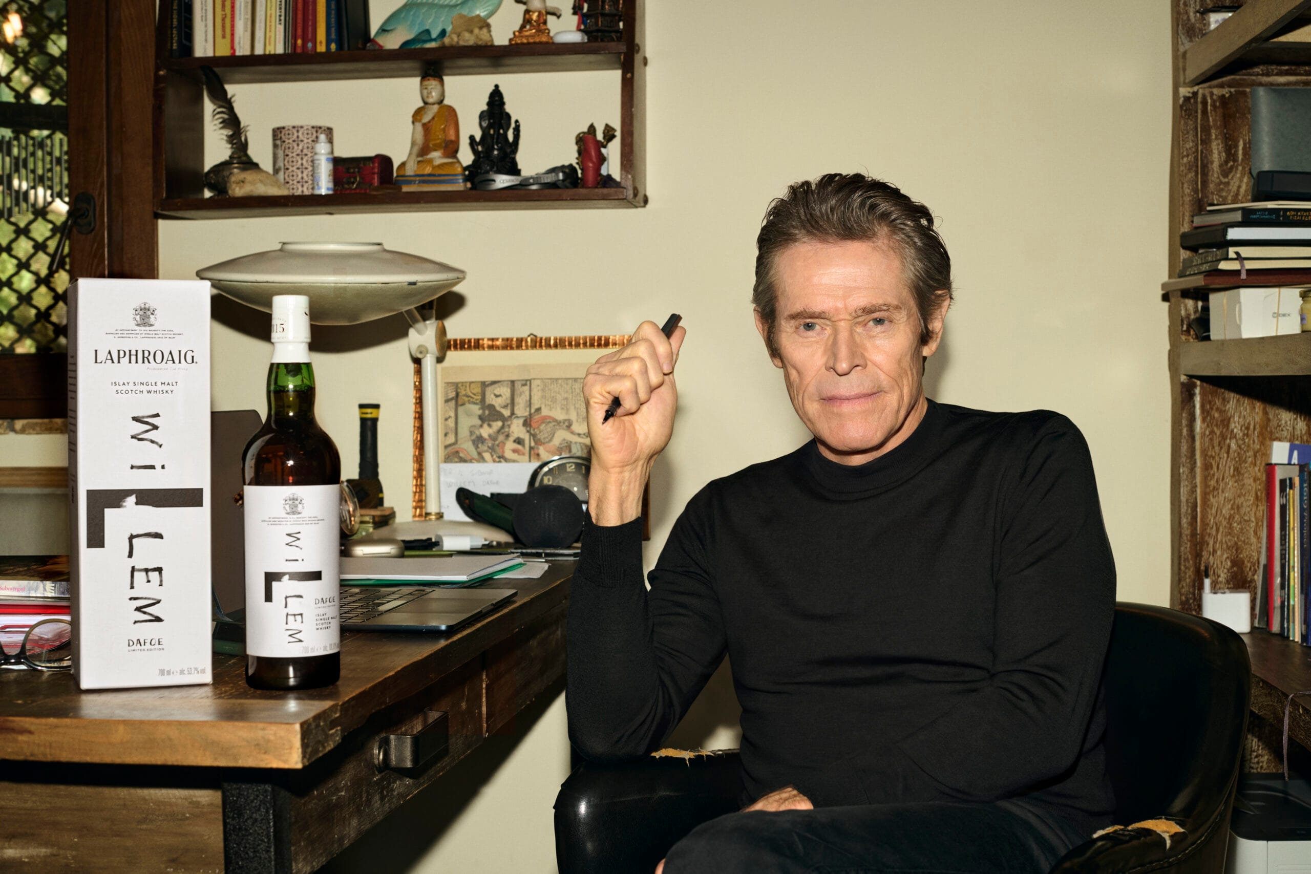 Willem Dafoe zit aan een bureau met een fles en verpakking van Laphroaig Willem Dafoe Limited Edition whisky