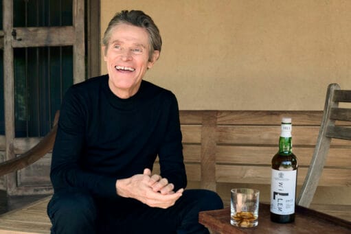 Willem Dafoe lachend op een houten bank naast een fles Willem Dafoe Islay Single Malt Scotch Whisky
