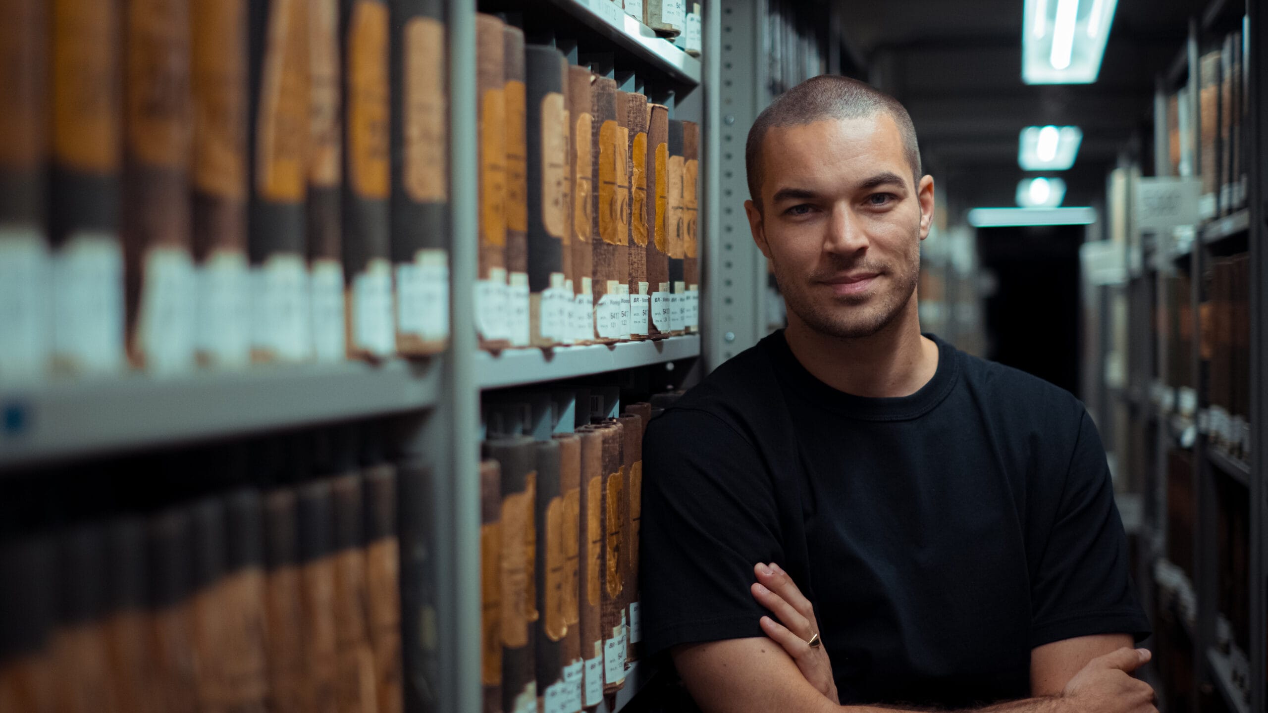 Een glimlachende man met kortgeschoren haar staat met de armen over elkaar in een archief vol oude boeken