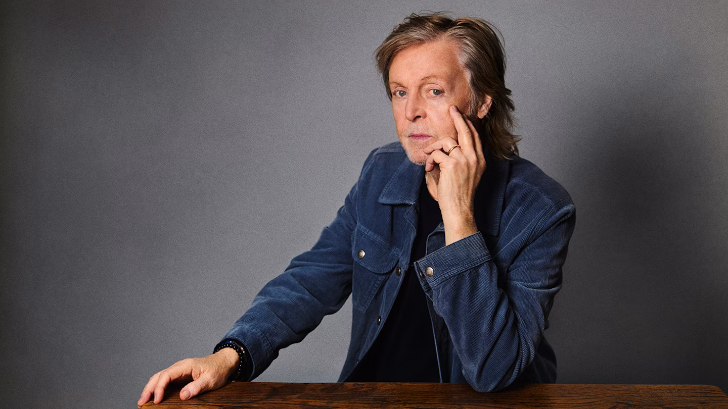 Portret van Paul McCartney in een blauw corduroy jasje, zittend aan een houten tafel tegen een grijze achtergrond