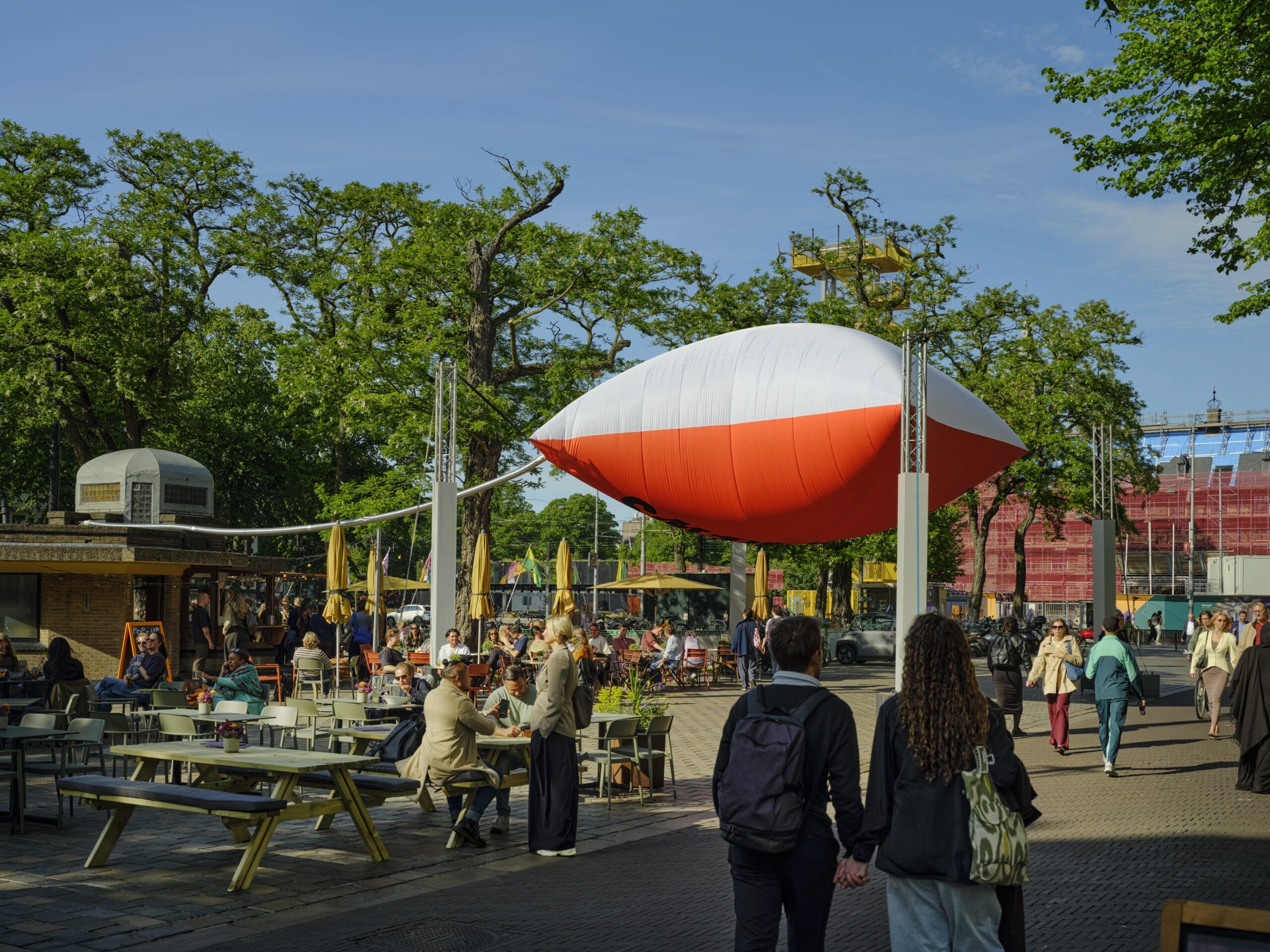 Mensen op een zonnig terras bij het Museumpark in Rotterdam, met een grote rood-witte opblaasbare vorm boven de tafels