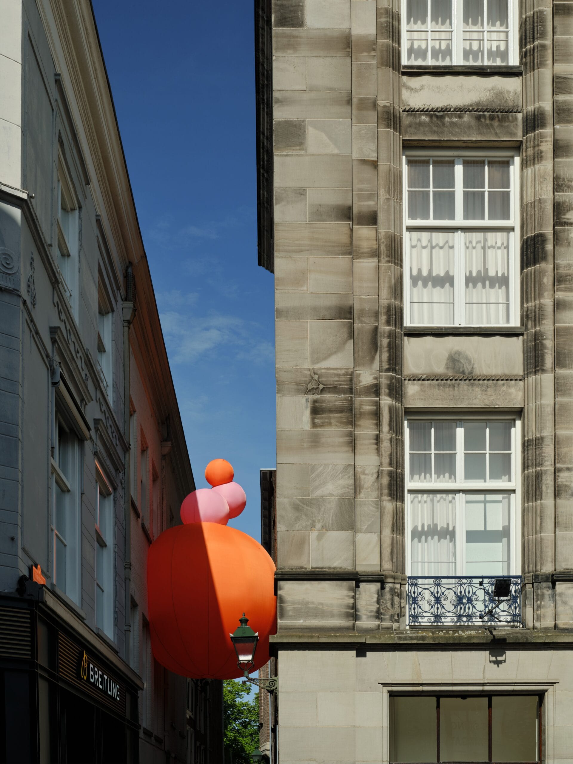 Grote oranje en roze opblaasbare bollen die tussen twee historische gebouwen in een stadsstraat hangen