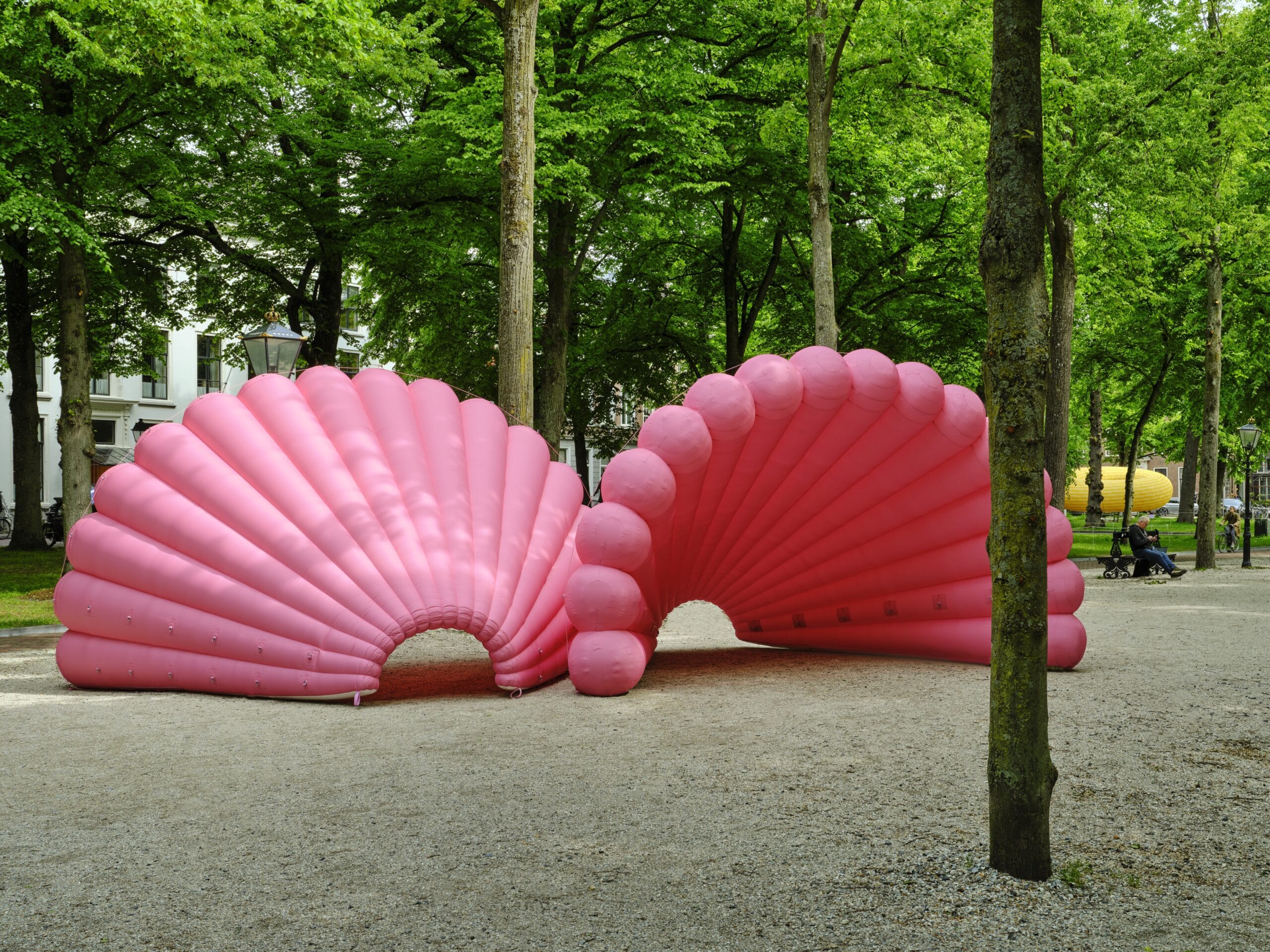 Twee grote, roze opblaasbare sculpturen in de vorm van schelpen op een grindpad in een park met groene bomen