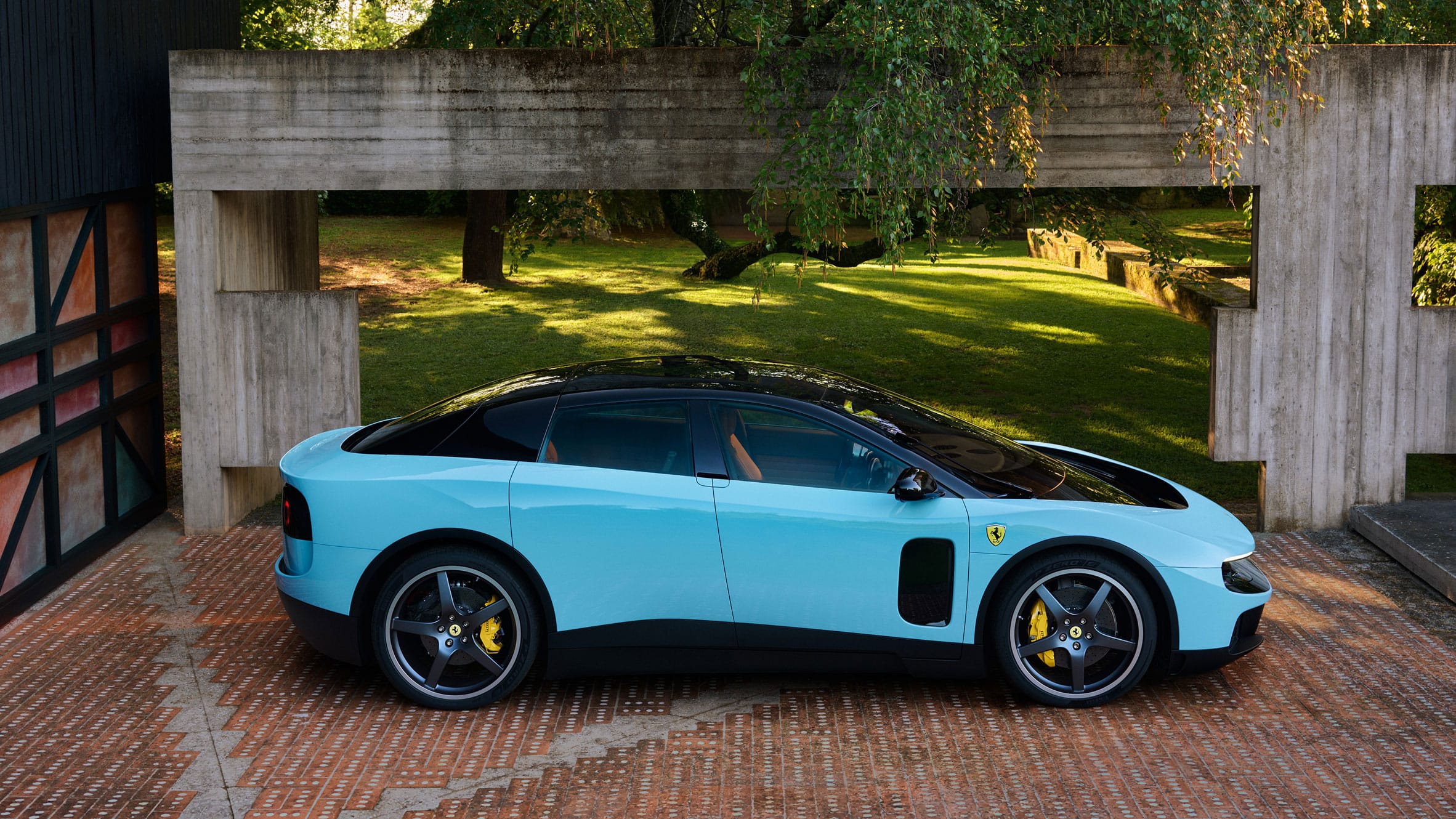 Zijaanzicht van een lichtblauwe Ferrari Luce geparkeerd op een terras bij een moderne betonnen structuur