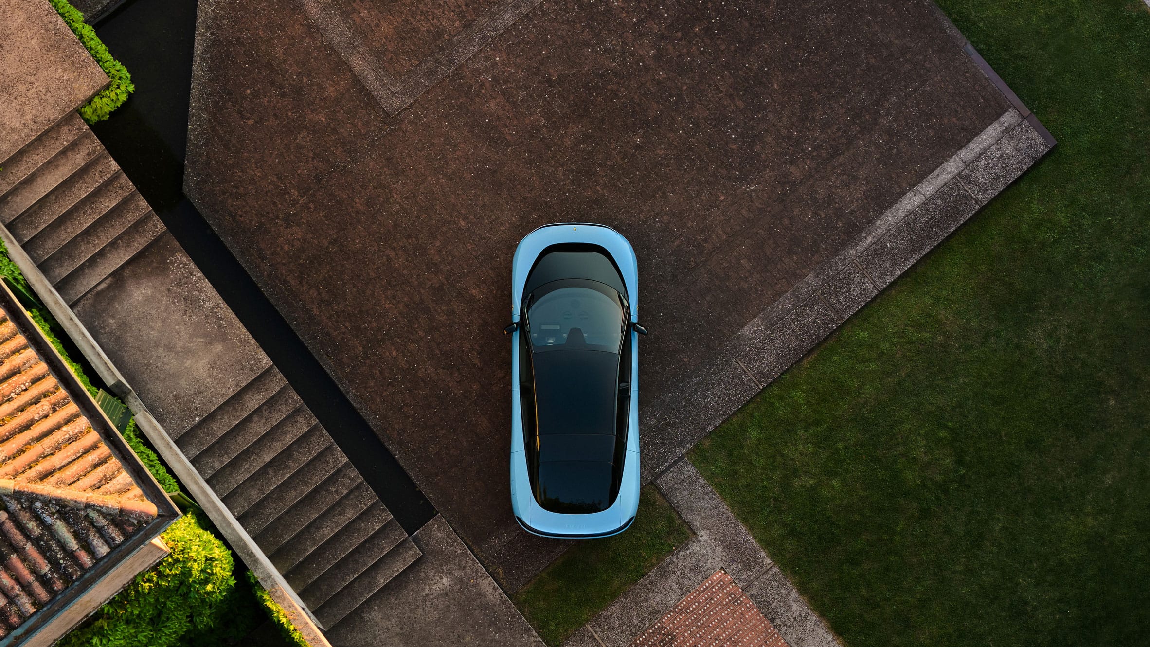 Bovenaanzicht van een lichtblauwe Ferrari Luce geparkeerd op een stenen oprit naast een gazon en een trap