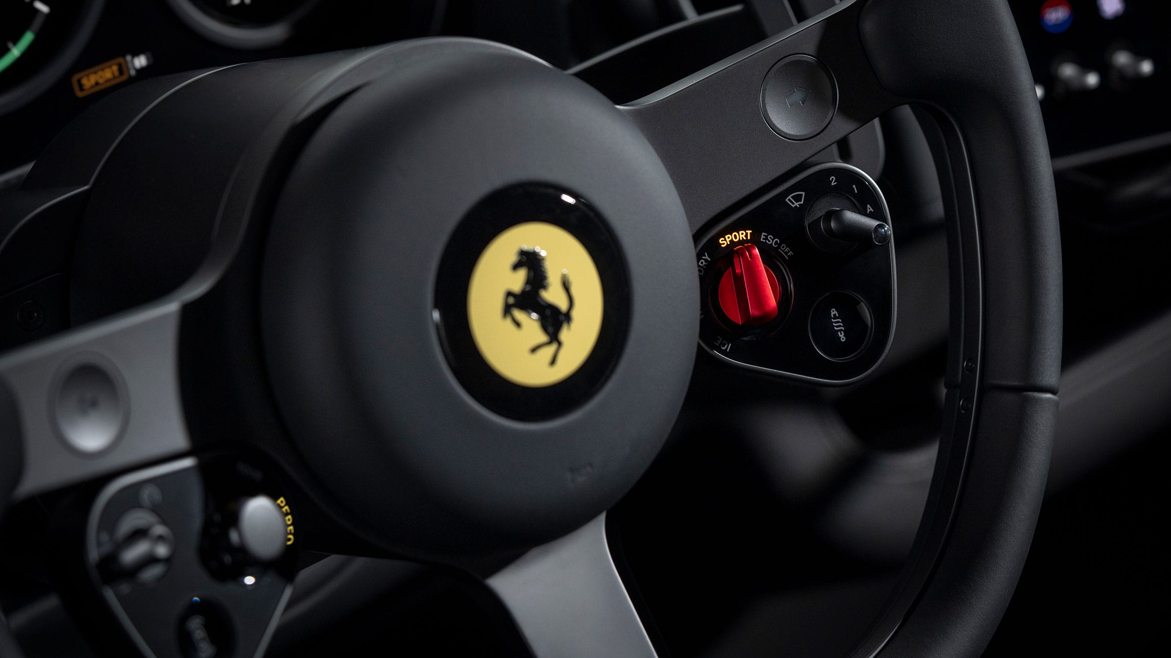 Detailopname van een Ferrari-stuurwiel met het gele logo en de rode Manettino-draaiknop ingesteld op de Sport-modus