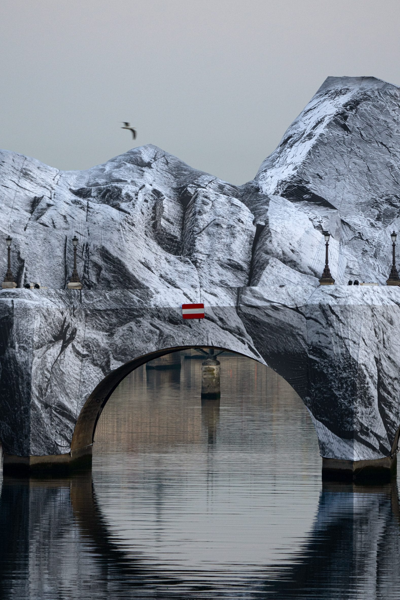 Een stenen brug over water die volledig is ingepakt in doeken met een zwart-wit print van besneeuwde bergen