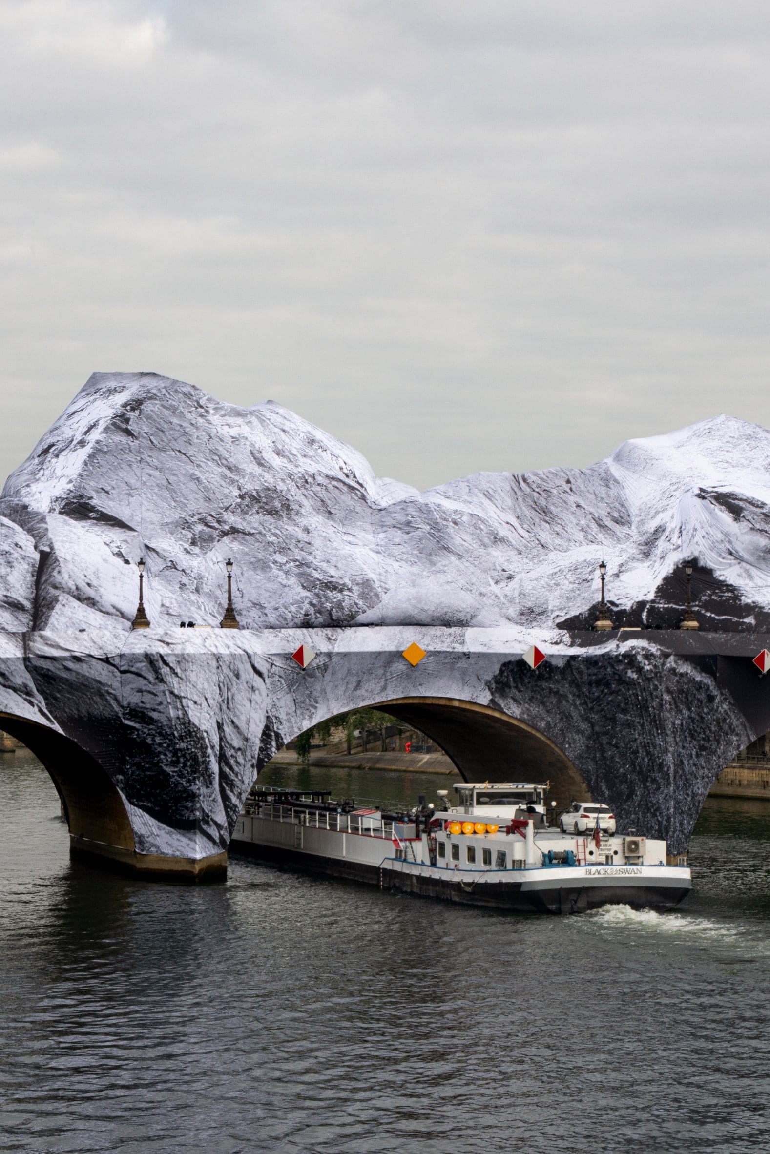 Een binnenschip vaart onder een Parijse brug die volledig is ingepakt in doeken met een afbeelding van besneeuwde bergen