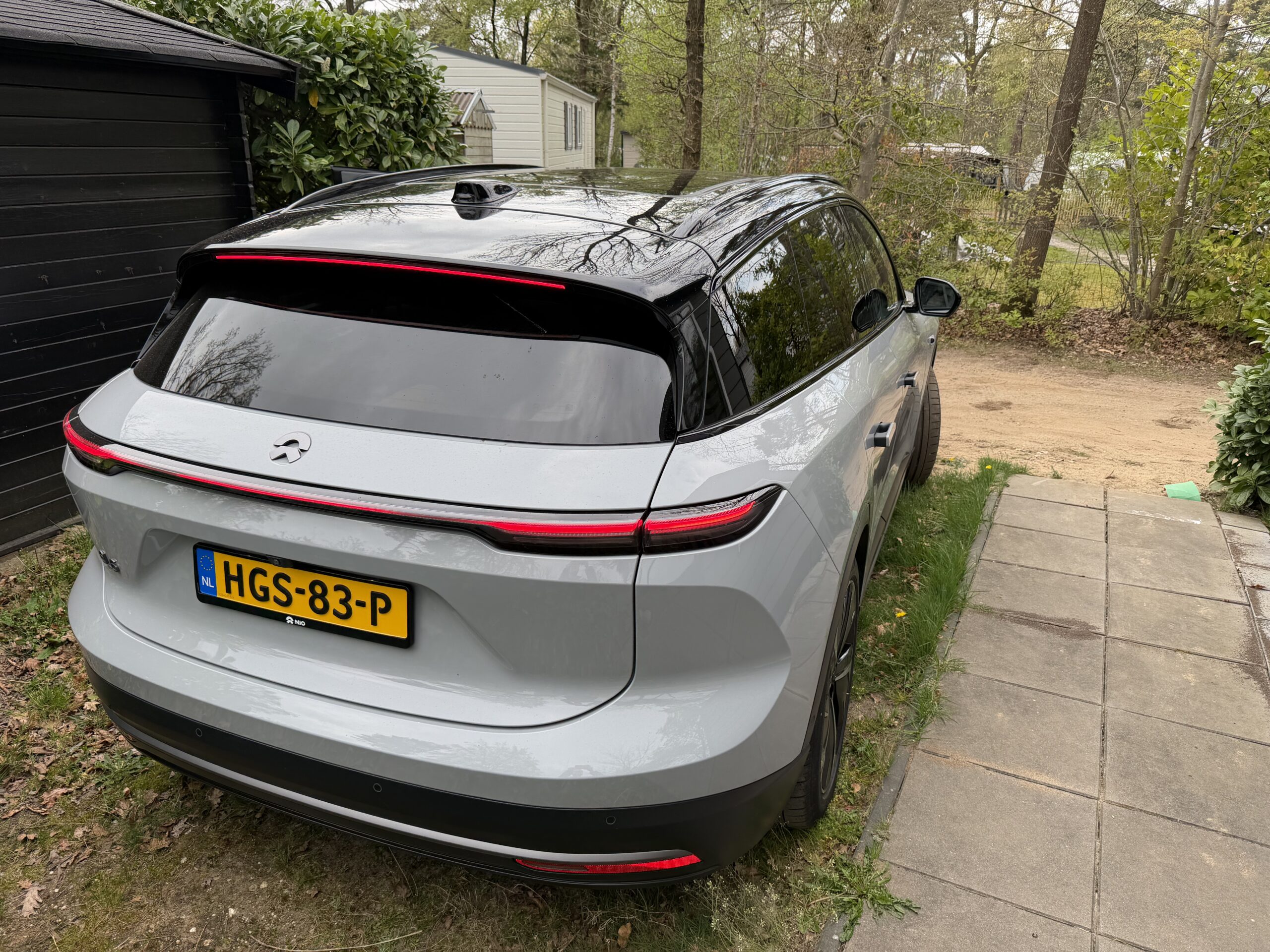 Achterzijde van een grijze Nio EL6 SUV met brandende achterlichten, geparkeerd op een oprit naast een houten schuur