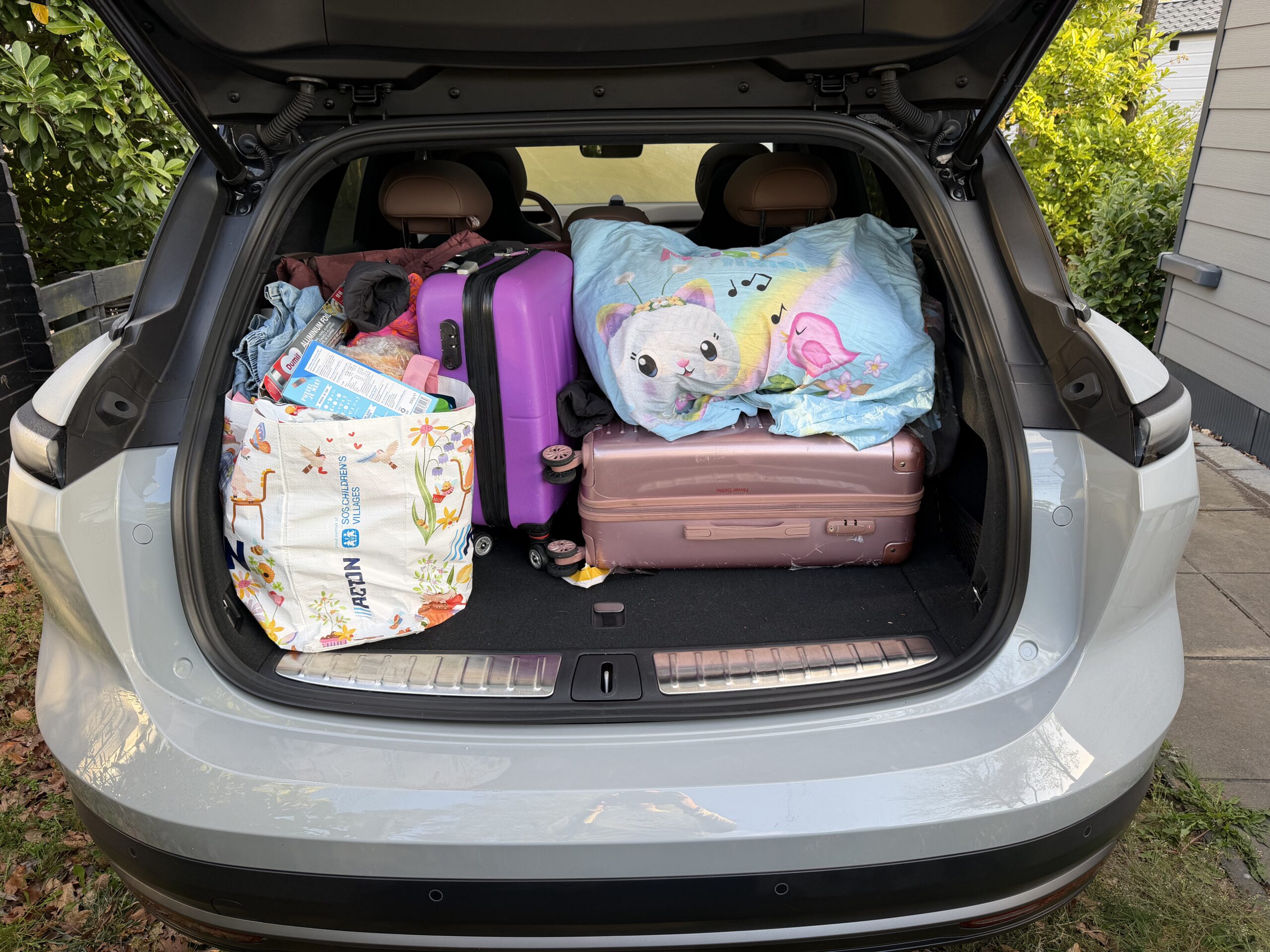 Een open kofferbak van een auto gevuld met koffers, een boodschappentas en een kussen met een kinderprint