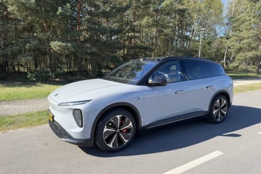 Een lichtgrijze NIO EL6 SUV geparkeerd op een asfaltweg met een bosrijke achtergrond op een zonnige dag