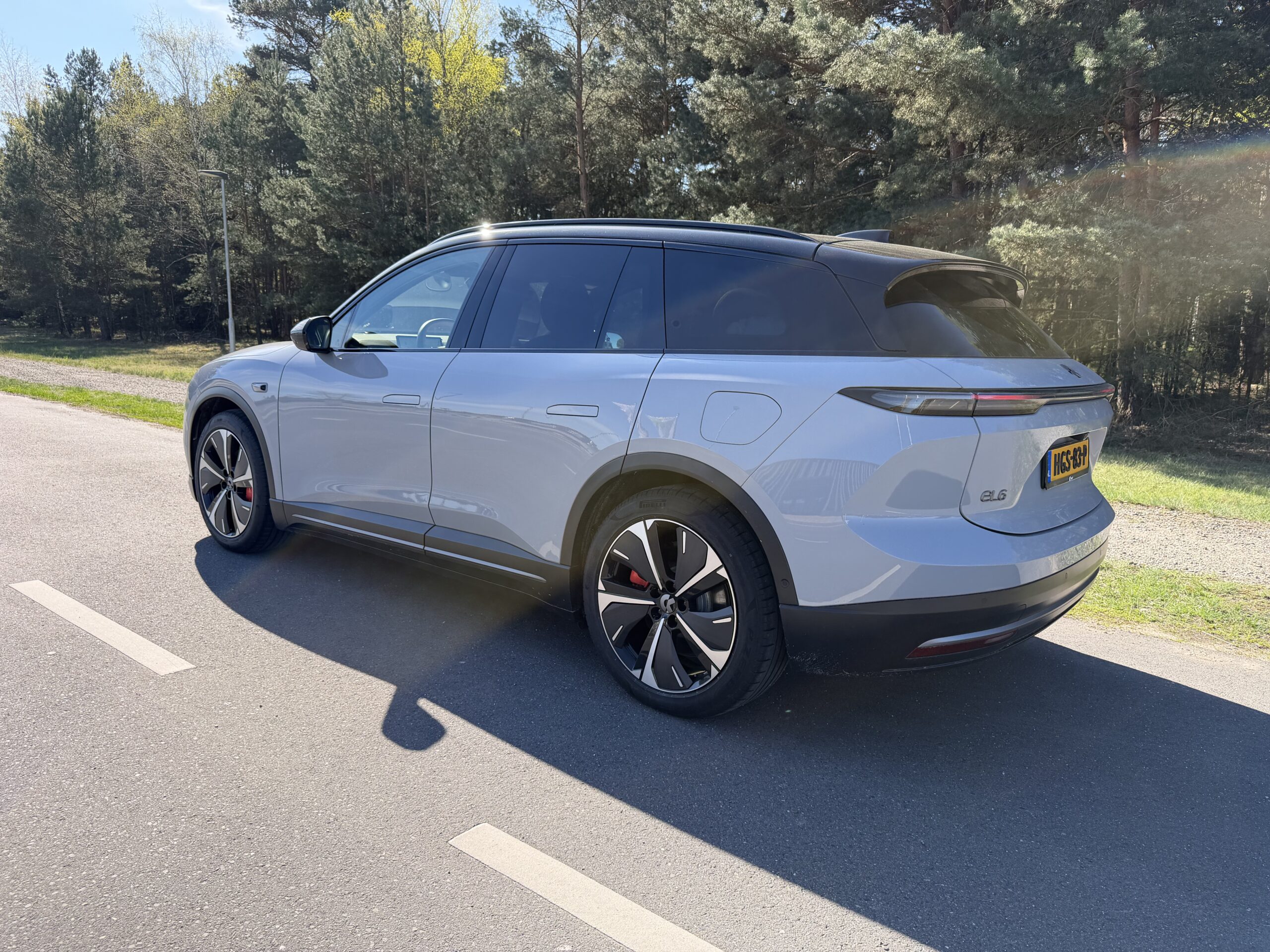 Zijaanzicht van een lichtgrijze Nio EL6 SUV geparkeerd op een weg langs een bosrand