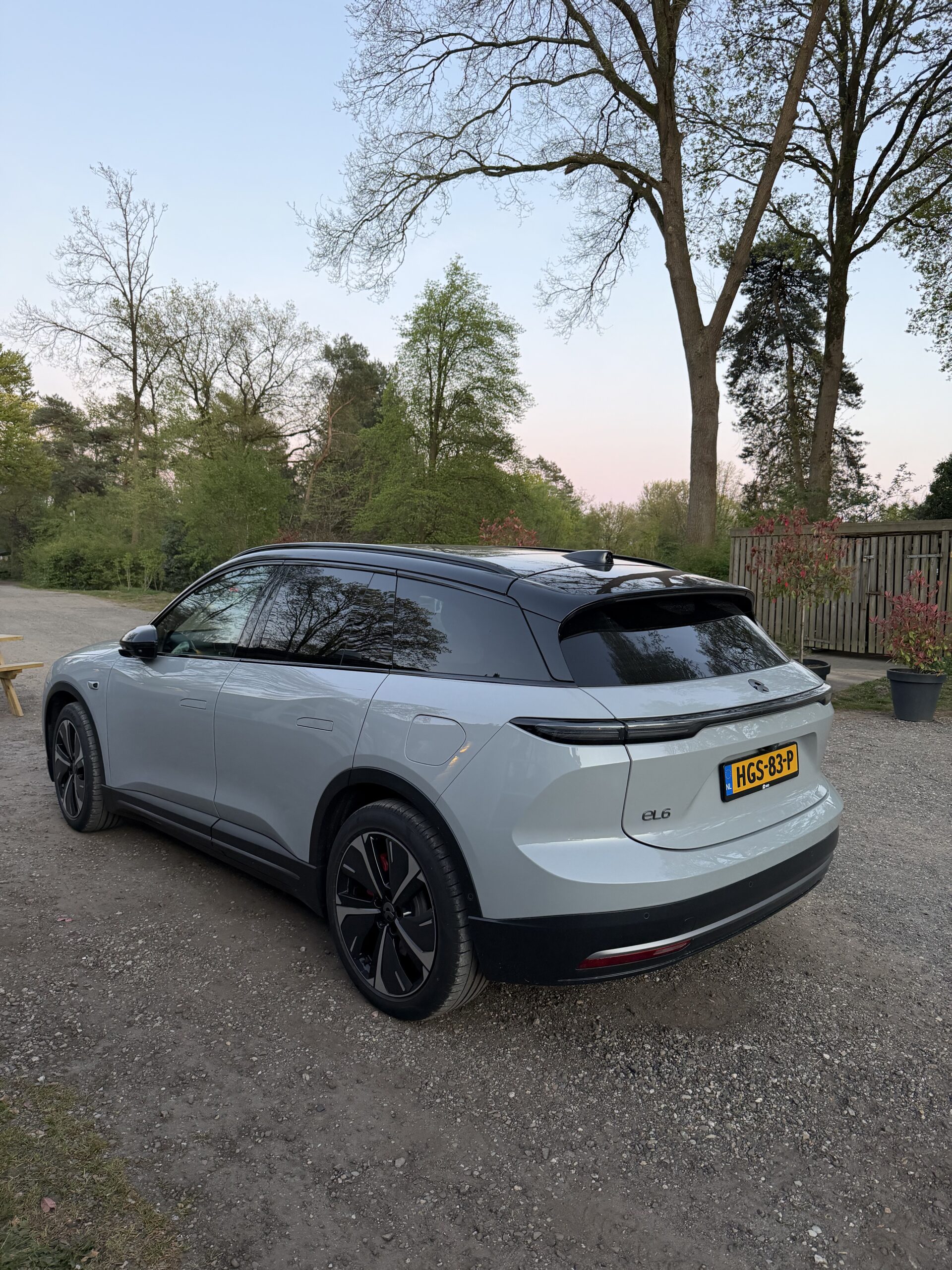 Zijaanzicht van een lichtgrijze NIO EL6 SUV geparkeerd op een grindpad in een bosrijke omgeving